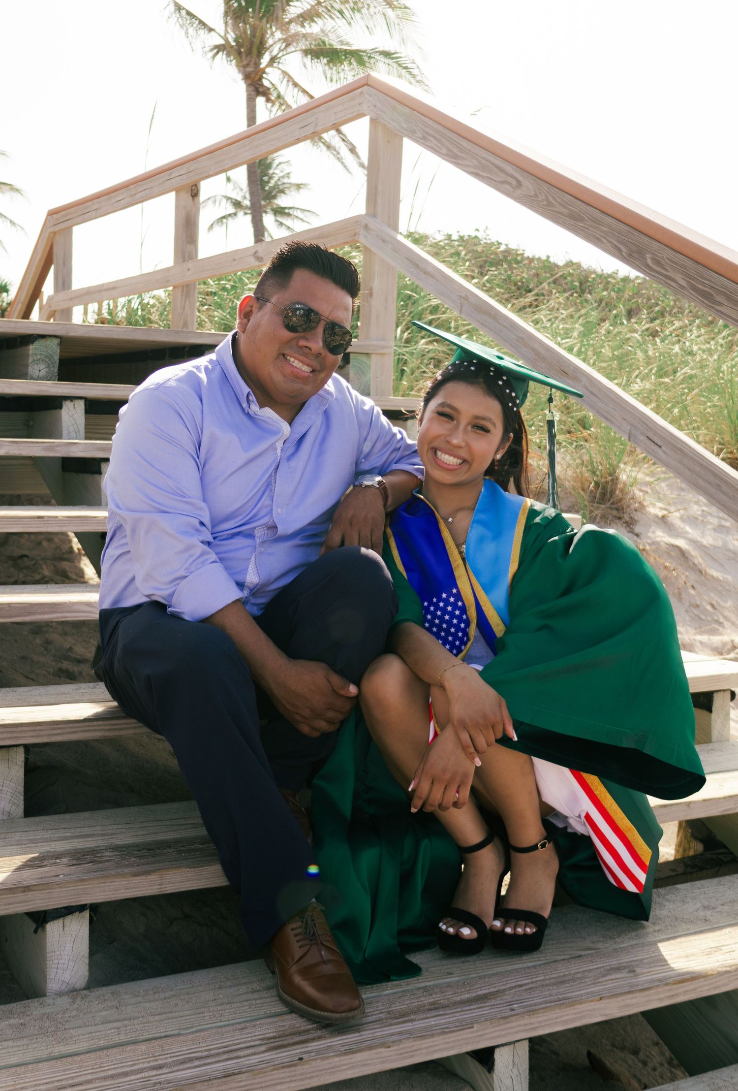Smiling graduate in green cap and gown poses with father on wooden beach boardwalk stairs.