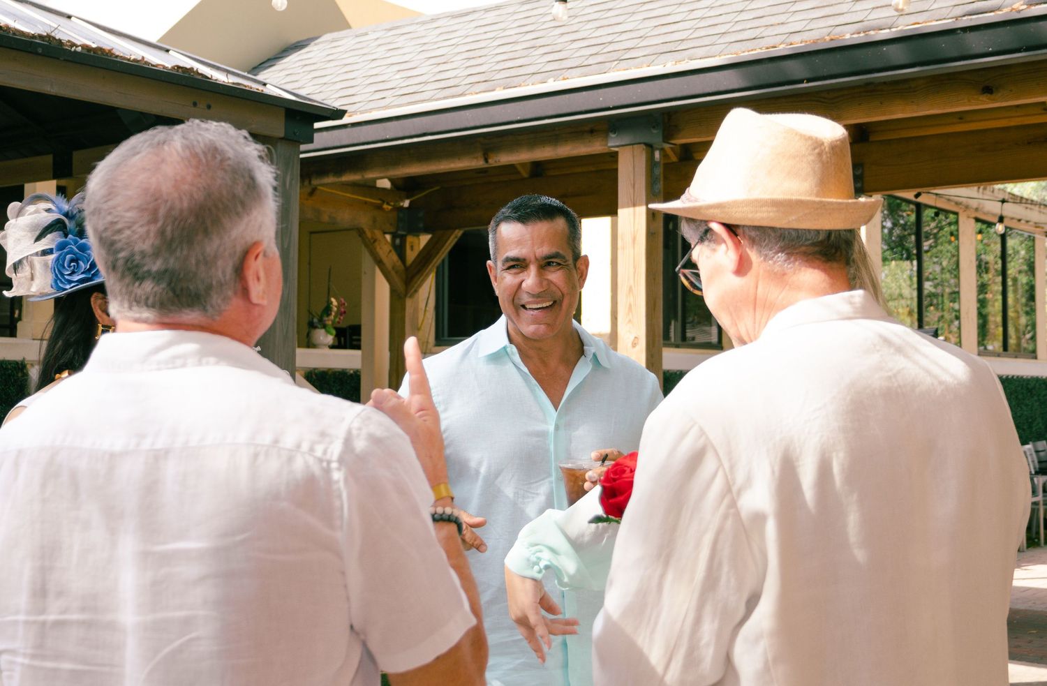 Three men socializing outdoors at a sunny garden party near a wooden pavilion.