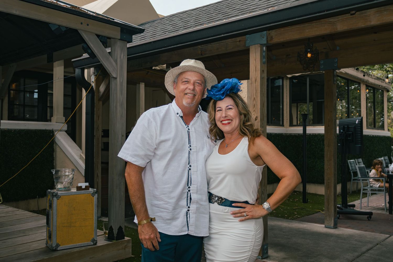 Couple posing outdoors, man in white shirt and hat, woman in white dress with blue fascinator.