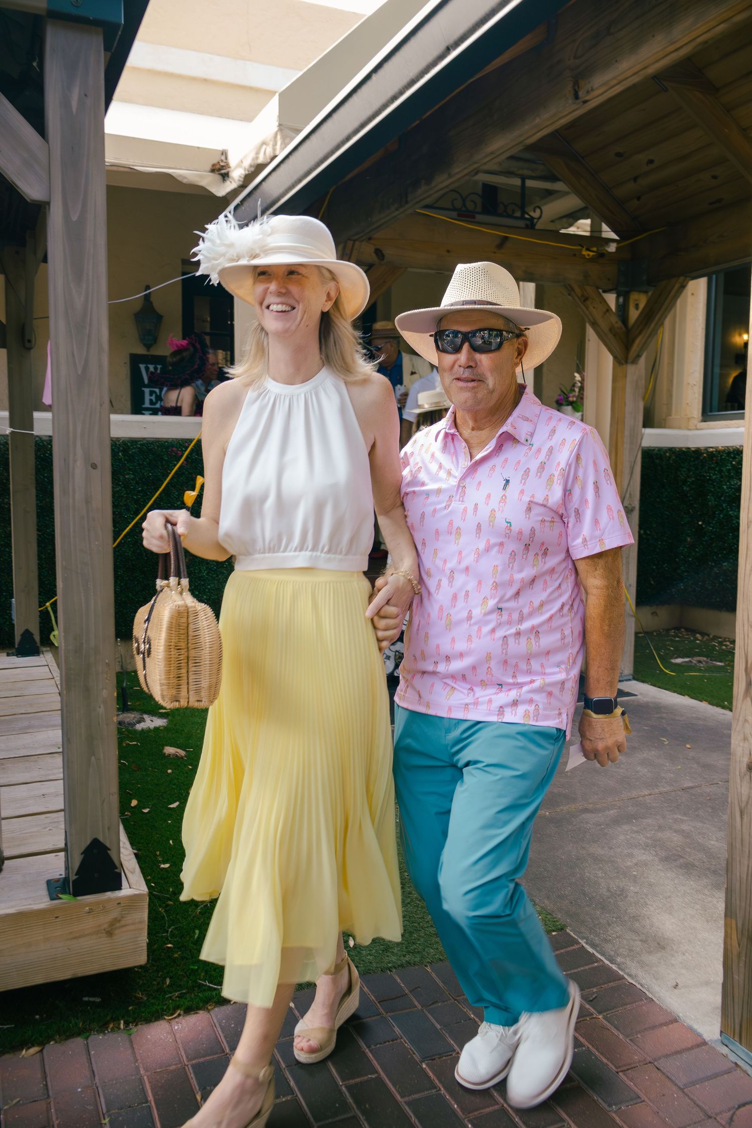 Couple in colorful Kentucky Derby attire with straw hats walk through outdoor venue.