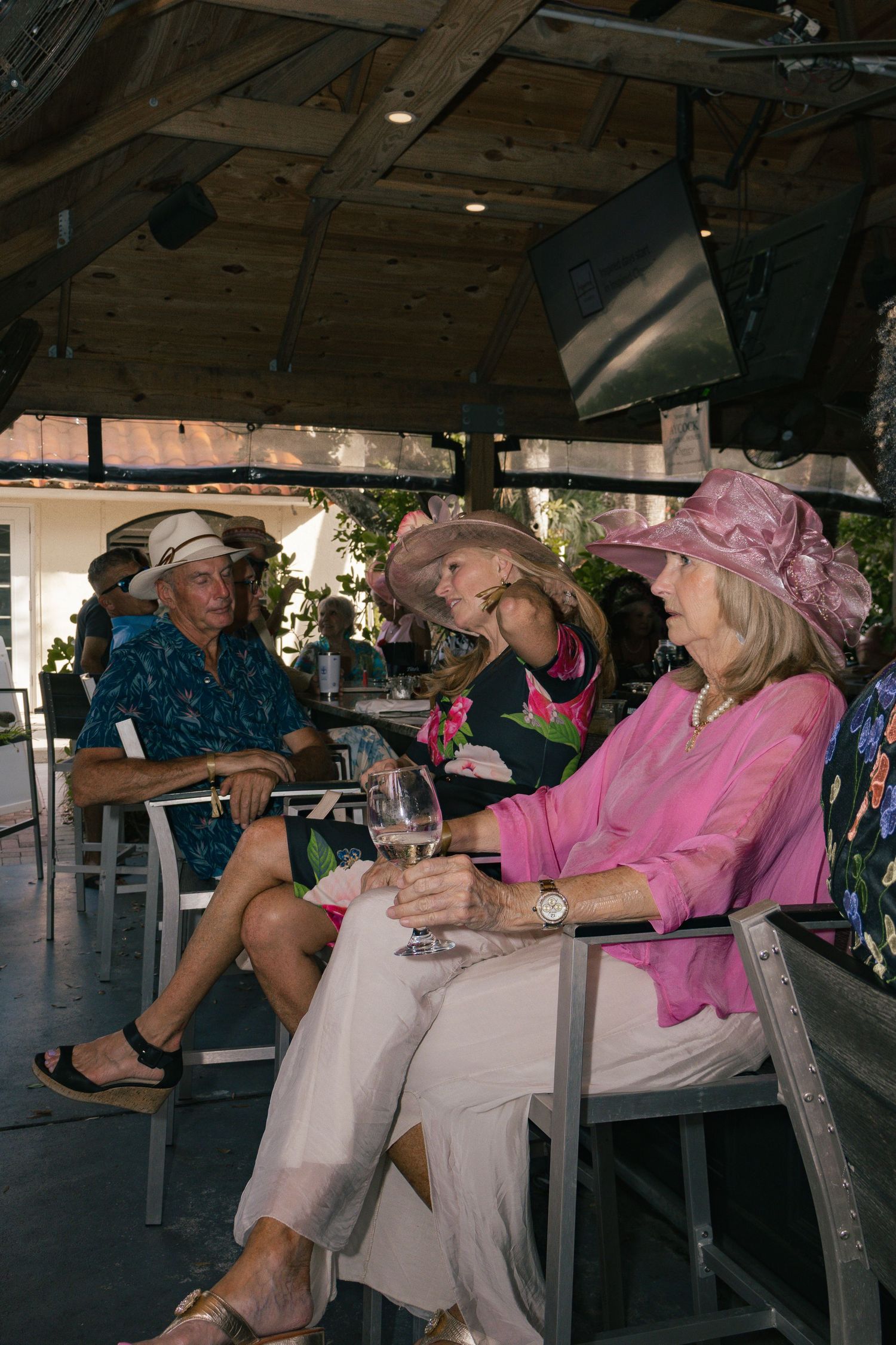 Elegant women in decorative hats enjoying wine at outdoor patio gathering.