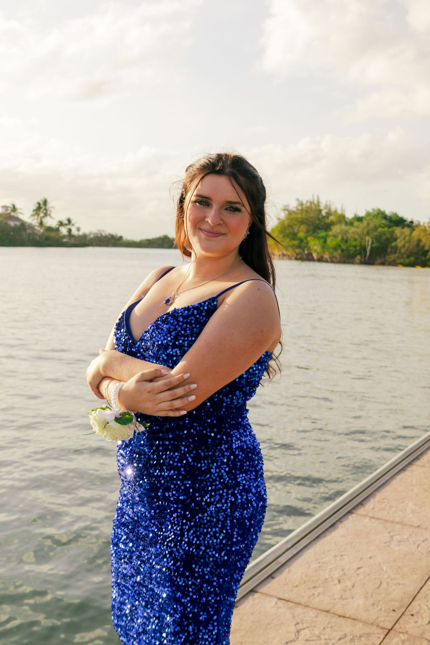 A smiling teenage girl in a royal blue sequin prom dress poses with arms crossed on a Florida waterfront.