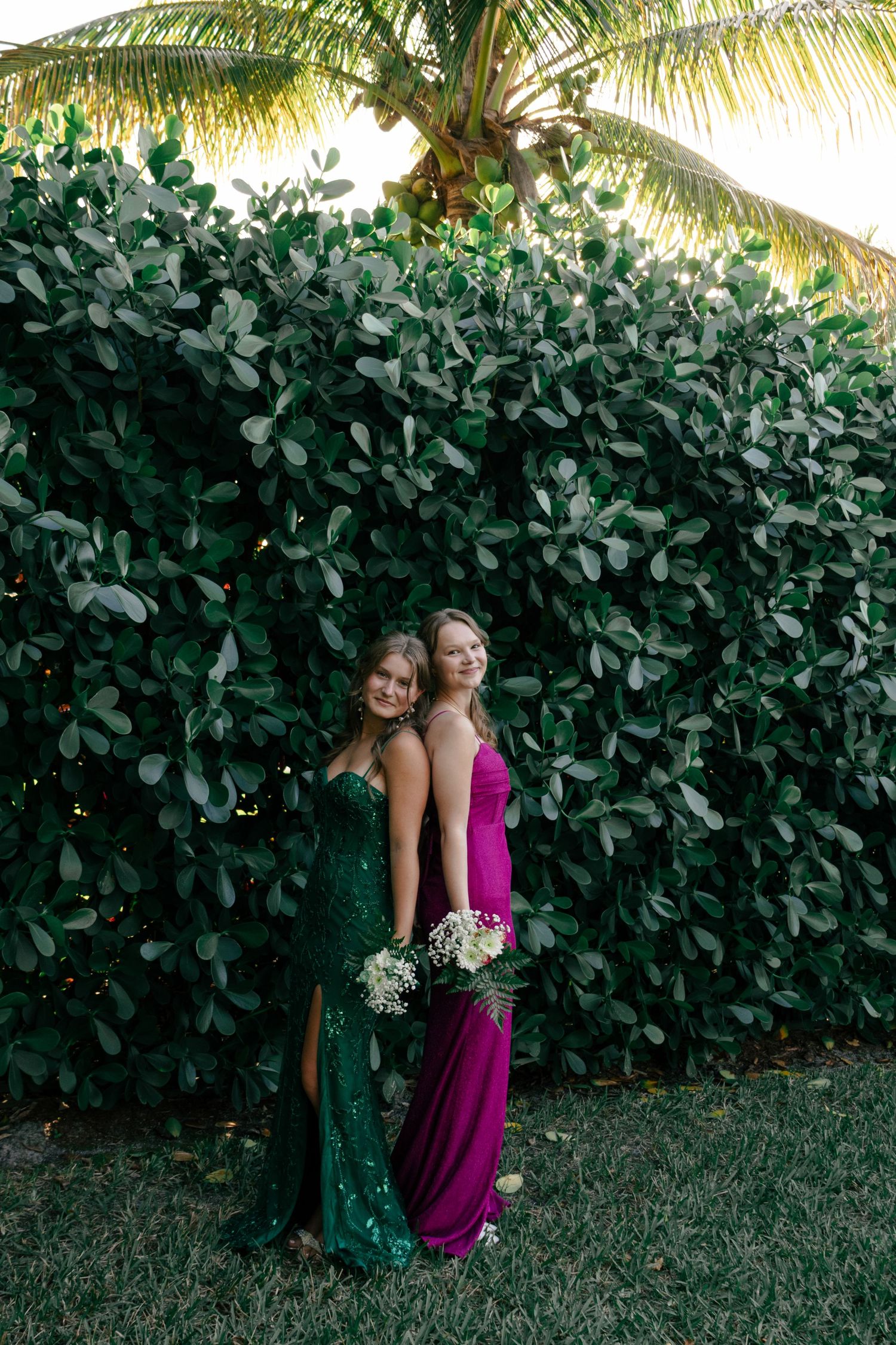 Two prom girls in green sequin and purple gowns pose back-to-back against a lush green hedge.