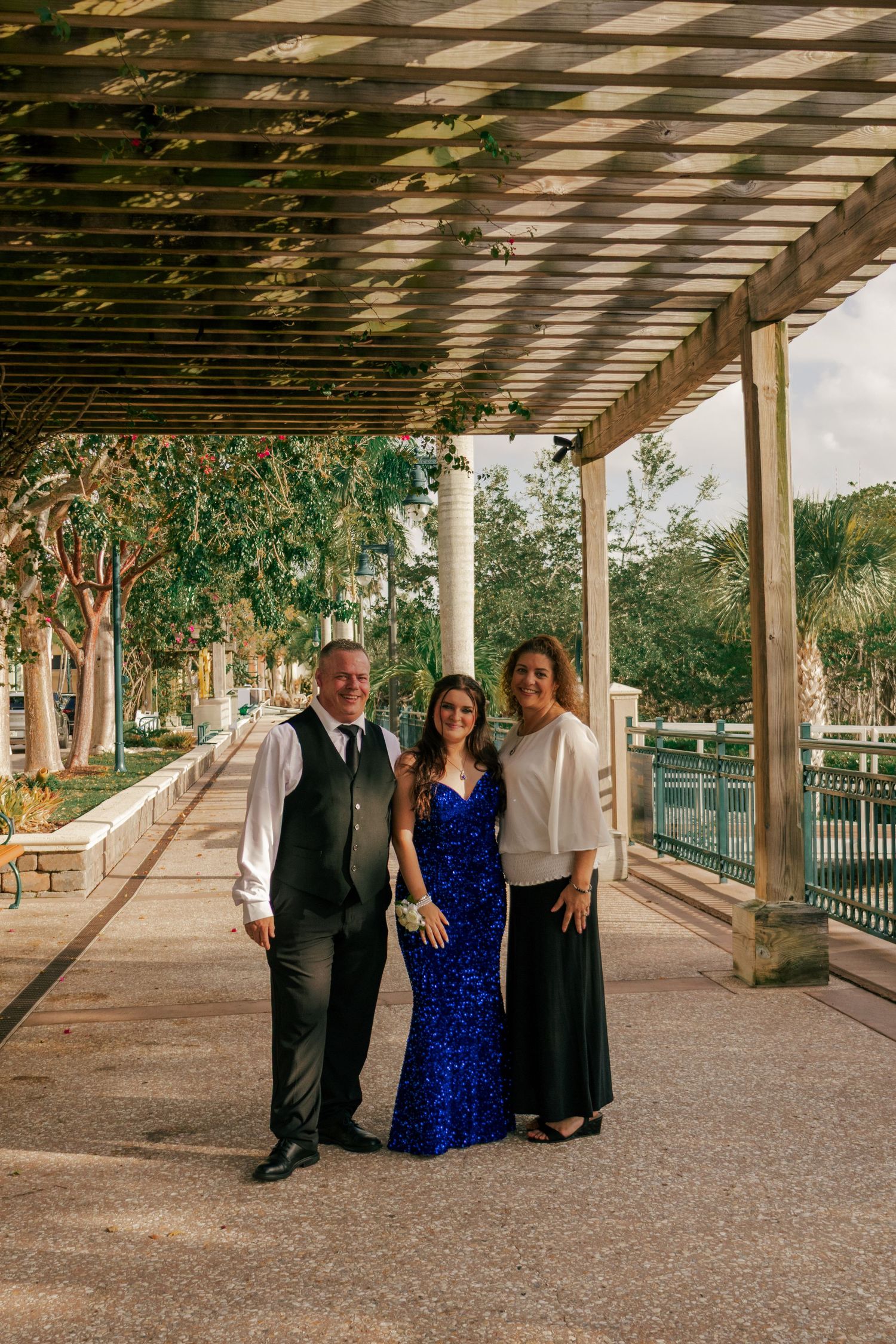 Family of three posing under pergola, girl in royal blue sequin gown flanked by parents in formal attire.