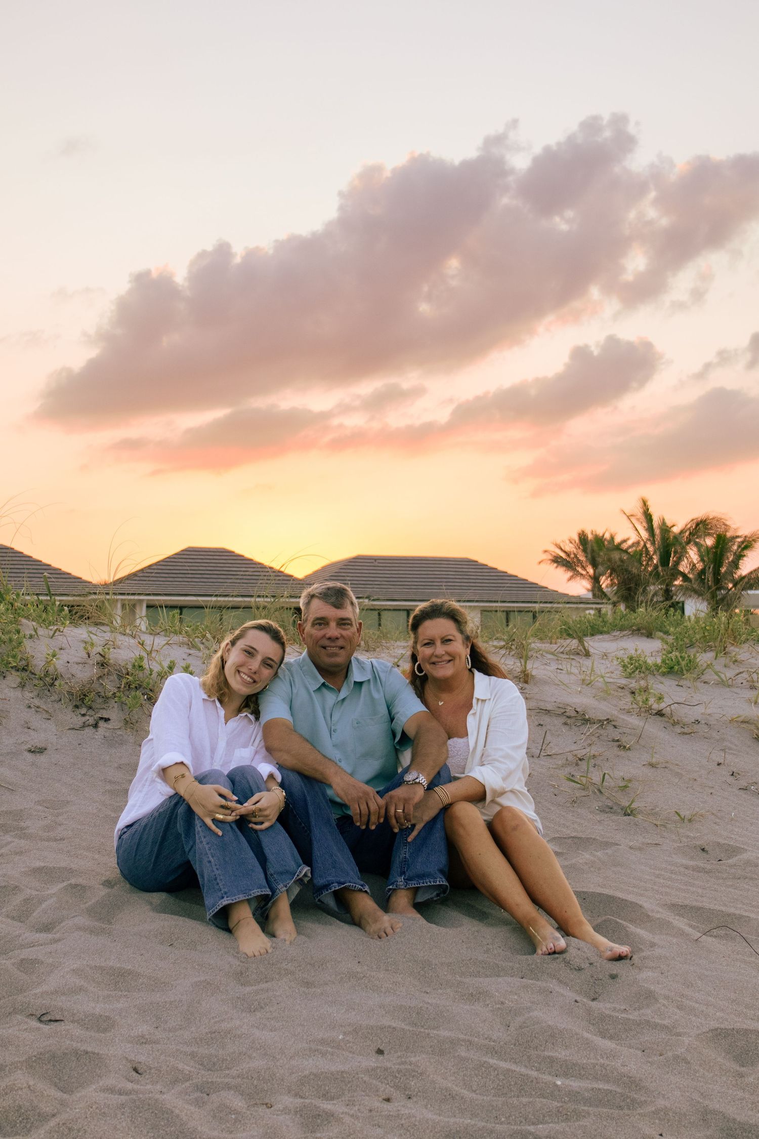 Family of three sitting on sandy beach at sunset with colorful sky and tropical backdrop.