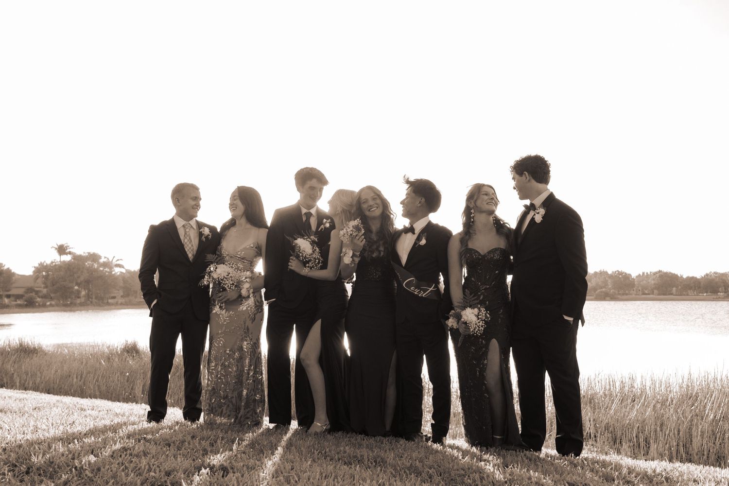 Group of elegantly dressed teens posing together near a lake for prom photos in sepia tone.