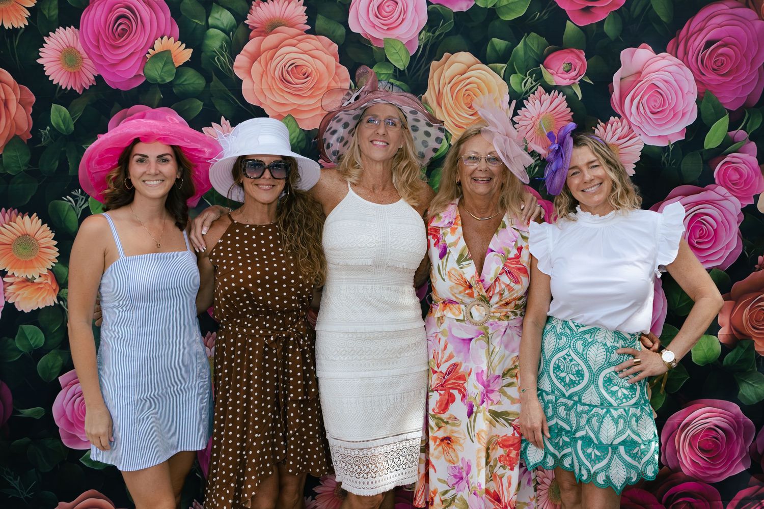 Five women wearing festive hats posing together in front of a bright floral rose backdrop.