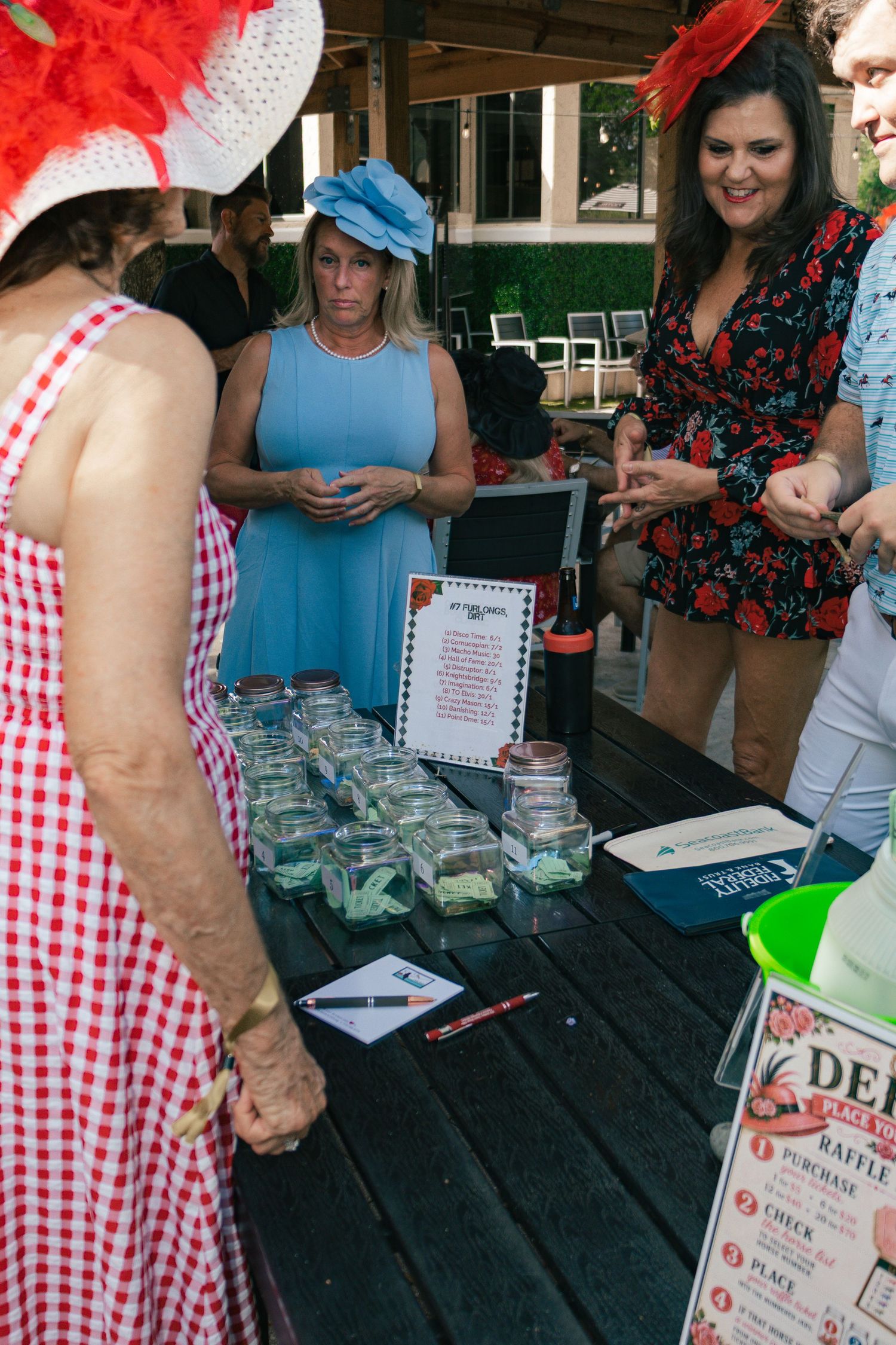 Women in festive hats browse jars at an outdoor raffle table during a Kentucky Derby themed party event.