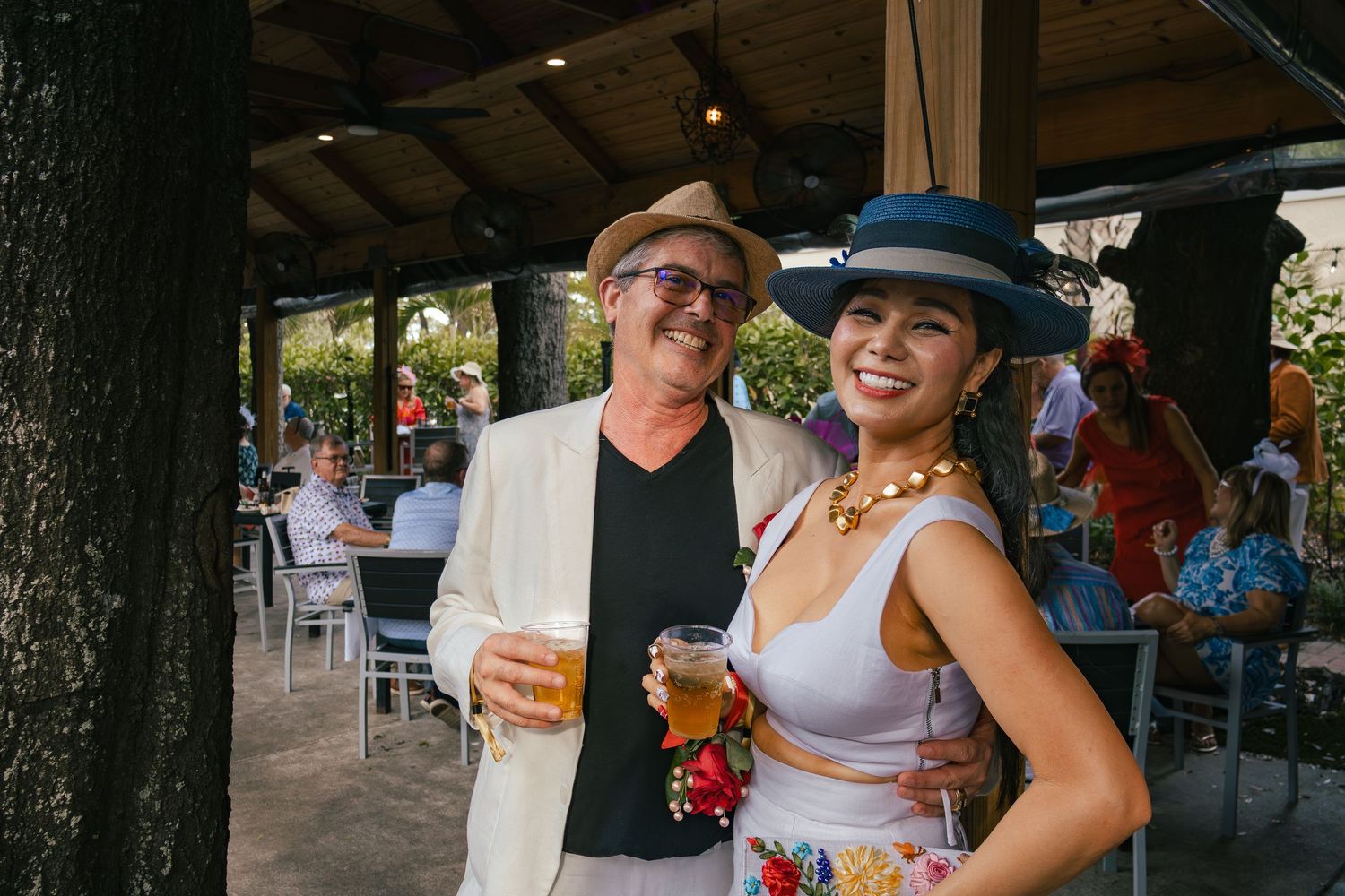 Man in tan suit and woman in floral dress with blue Derby hat pose together at outdoor party.