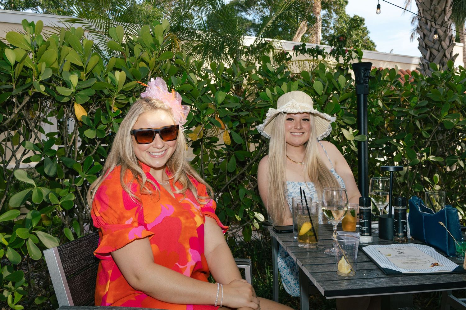 Two blonde women in colorful hats and dresses sitting at outdoor patio table with drinks.