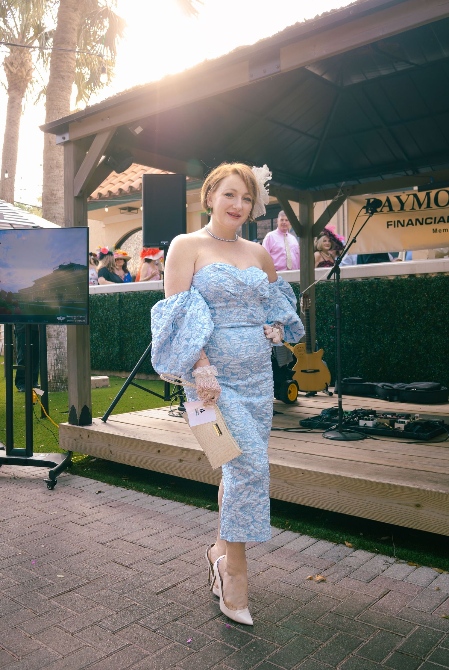 Woman in blue floral off-shoulder dress with puff sleeves poses at outdoor event near stage.
