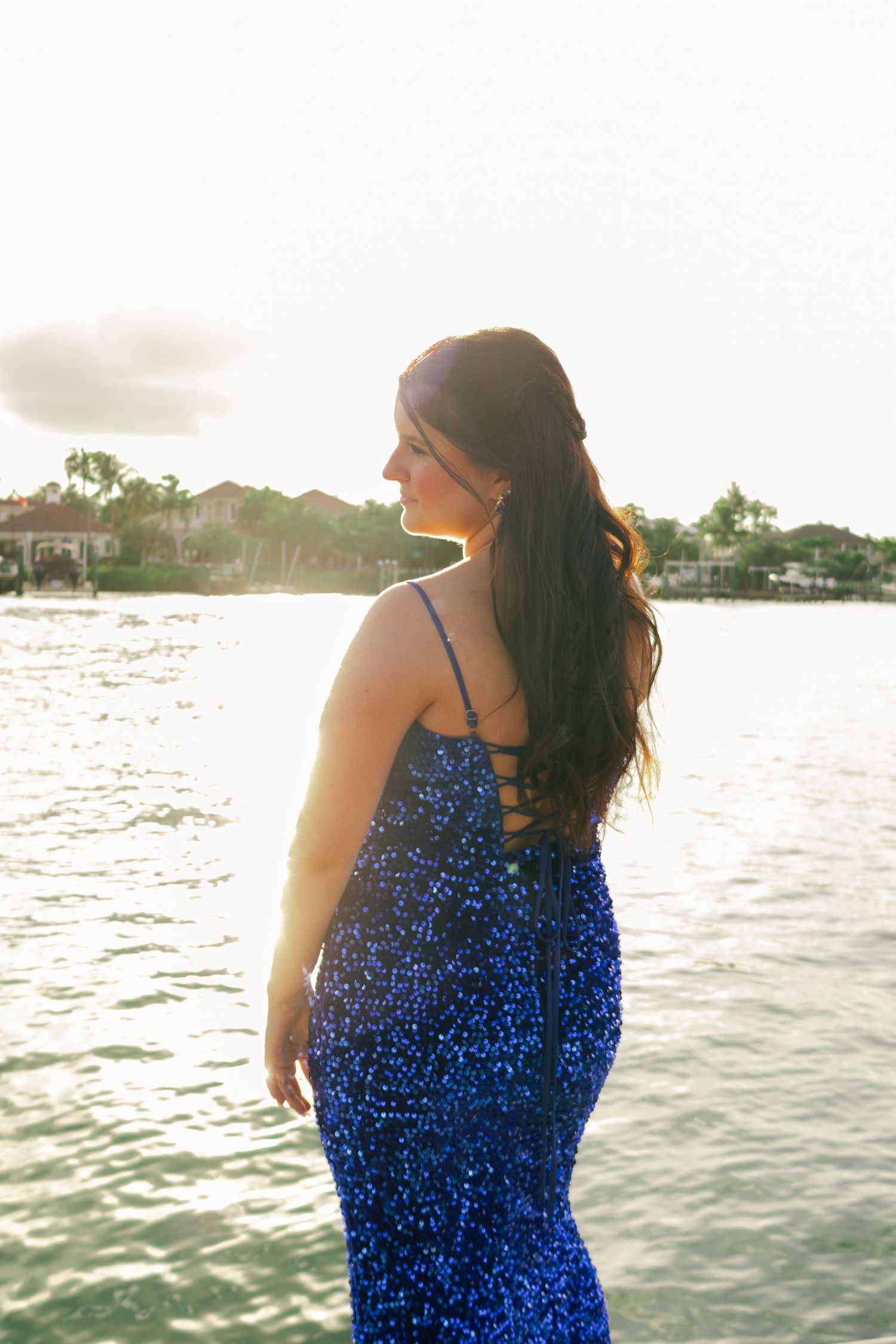 A teenage girl in a royal blue sequin mermaid prom dress poses backlit by golden sunset light over water.