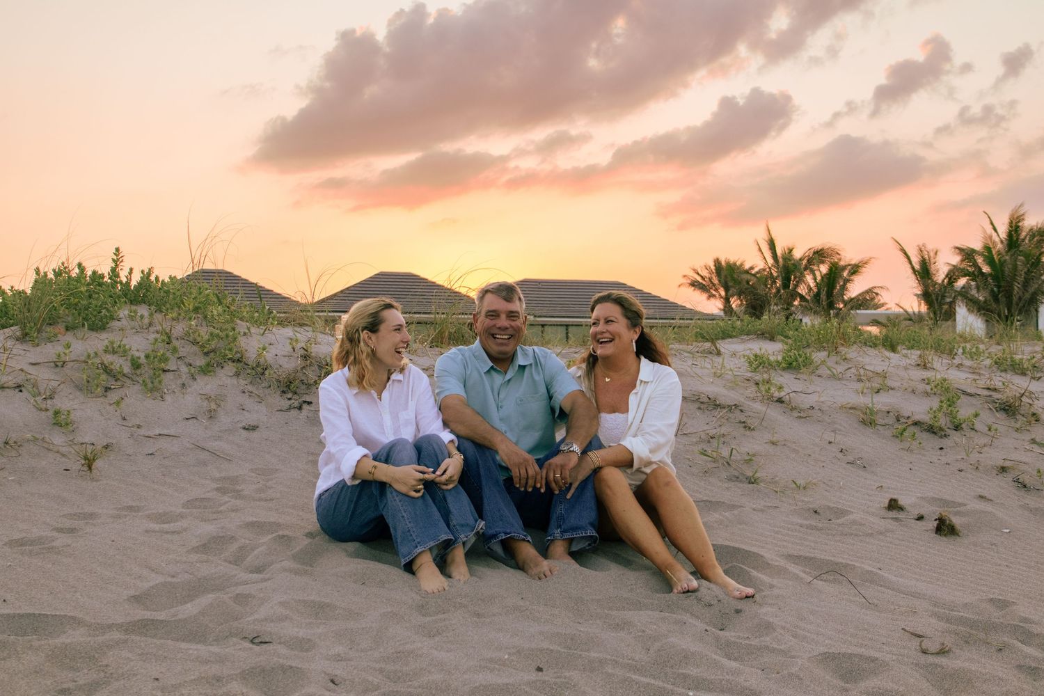 Three friends laughing together on a sandy beach at sunset with palm trees and colorful sky in the background.