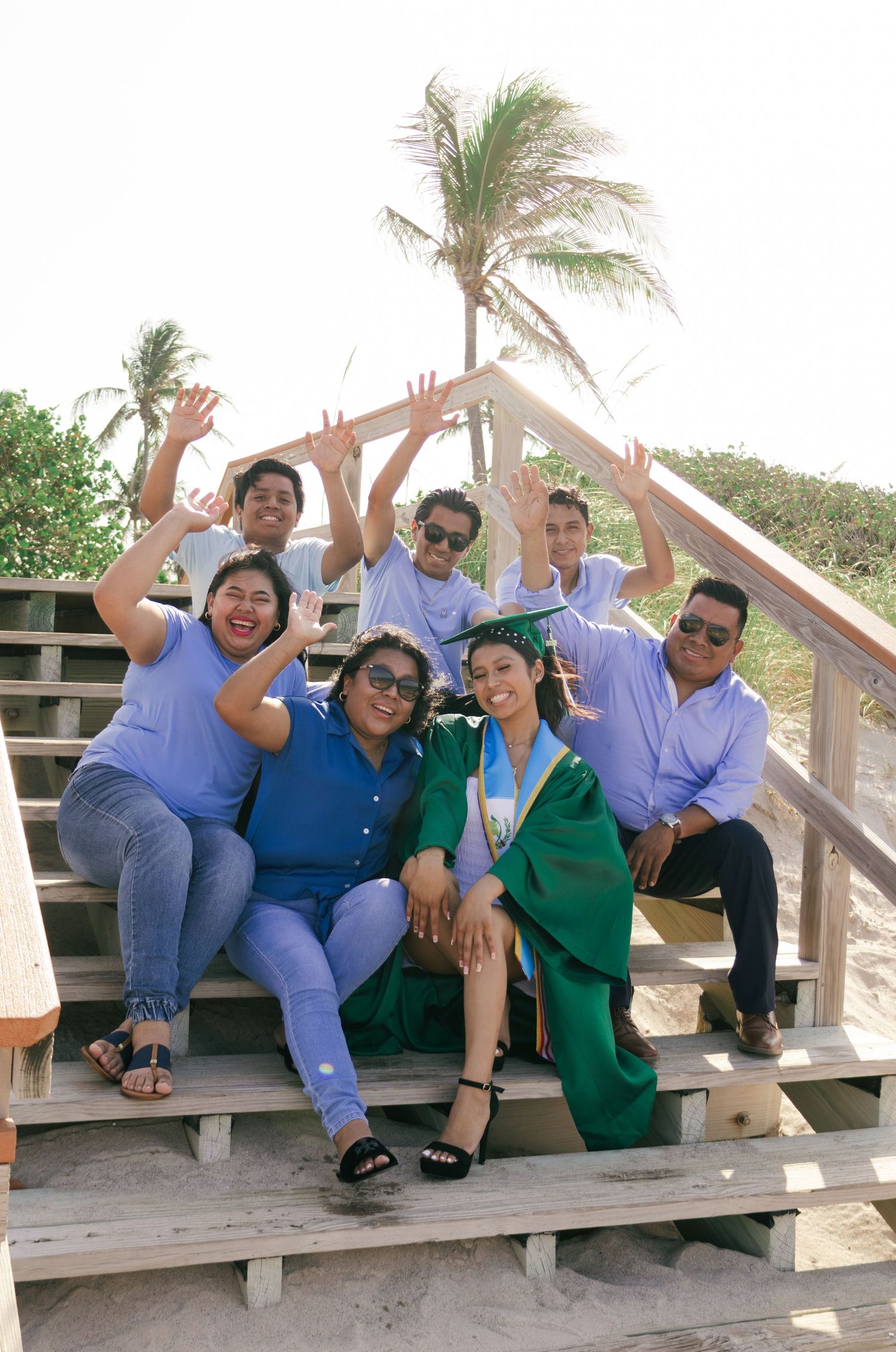 Group of friends celebrating a graduation at the beach, with the graduate in green cap and gown on wooden steps.