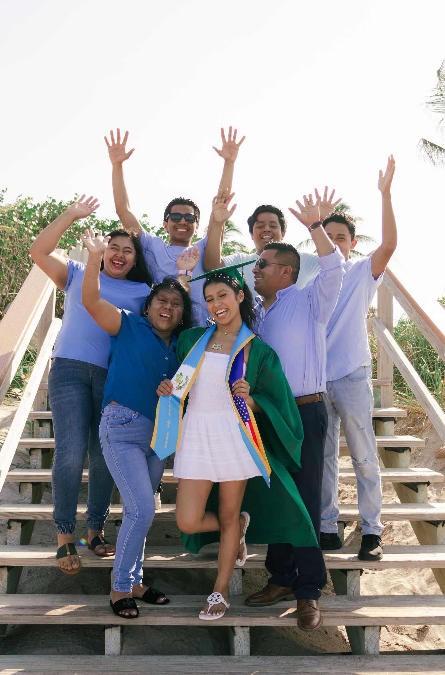 Joyful graduate in green gown celebrates with cheering family on sunny beach boardwalk steps.
