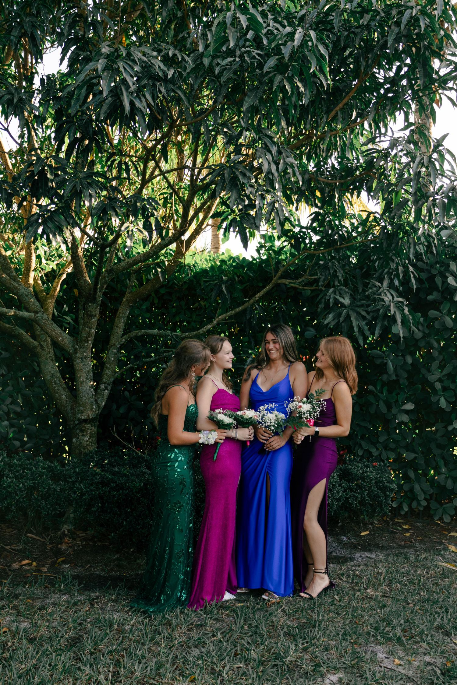 Four bridesmaids in colorful green, pink, blue, and purple gowns admiring bouquets under a lush tree.