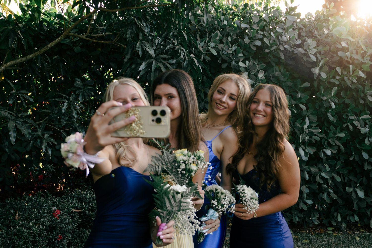 Four bridesmaids in blue dresses taking selfie with bouquets in front of green hedge.