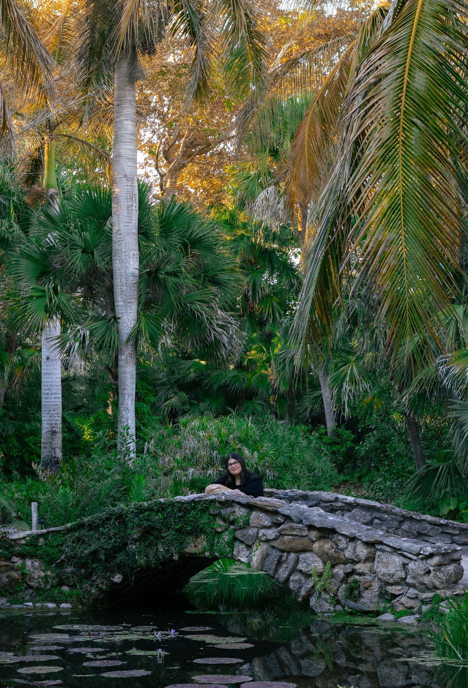 Young woman in black outfit sitting on stone bridge over pond in lush palm garden.