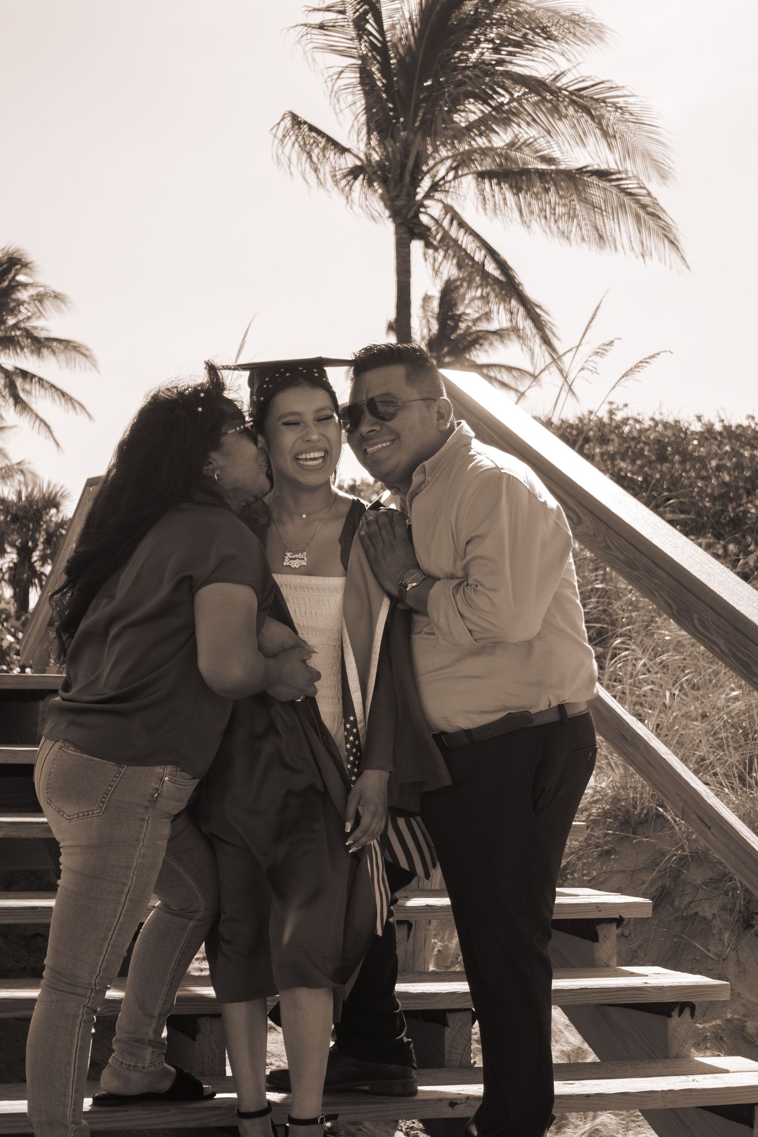 Sepia-toned photo of graduate in cap and gown laughing joyfully with two proud parents near palm trees.