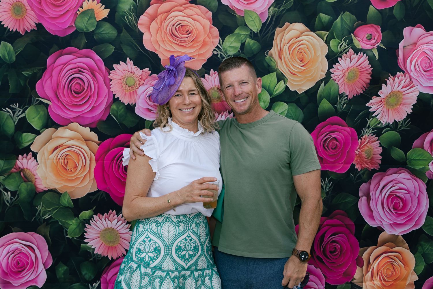 Couple smiling together, woman wearing purple fascinator hat, posing in front of colorful floral backdrop.