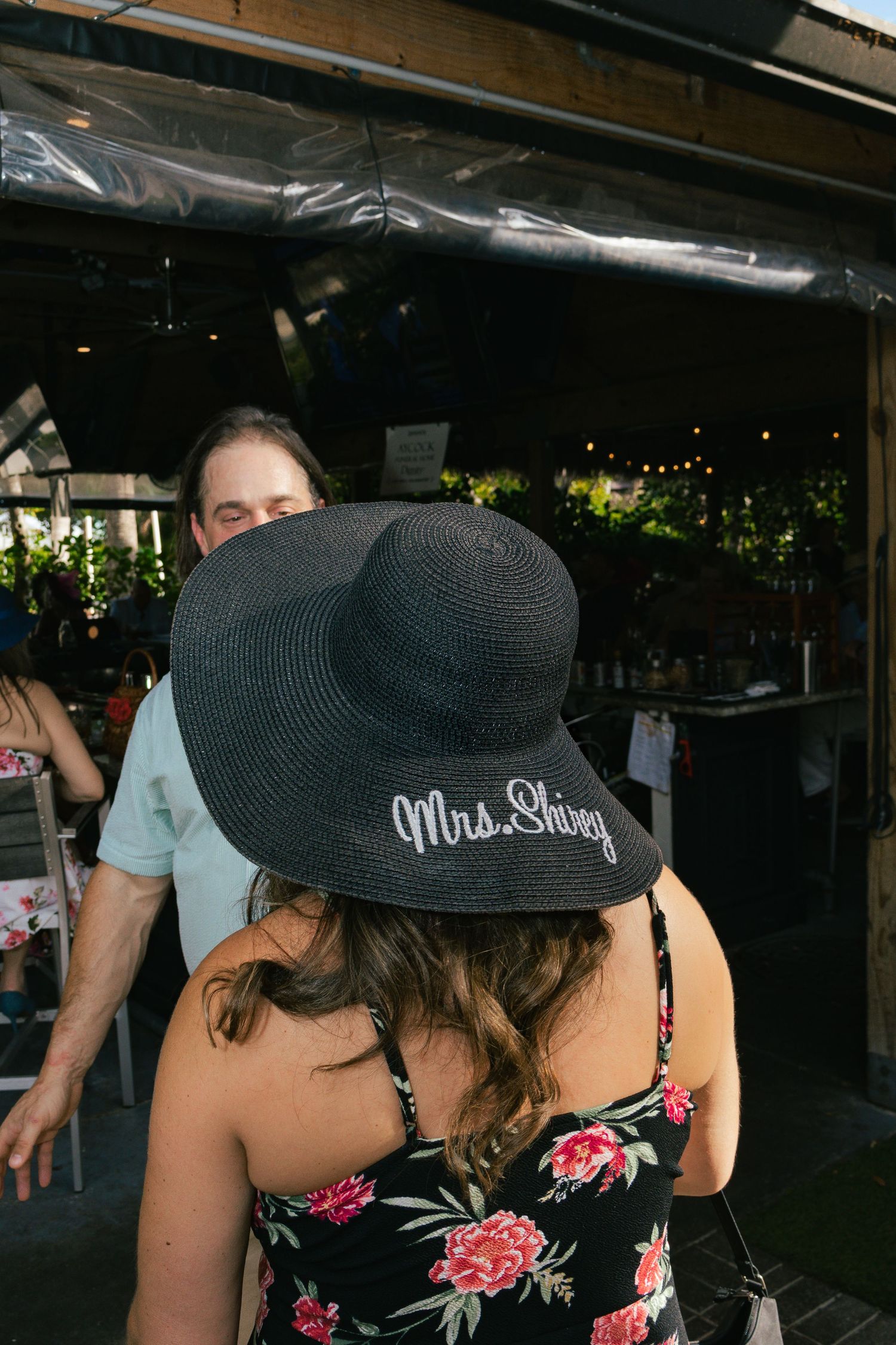 Woman wearing black floppy hat reading Mrs. Shirey at a festive outdoor restaurant event.