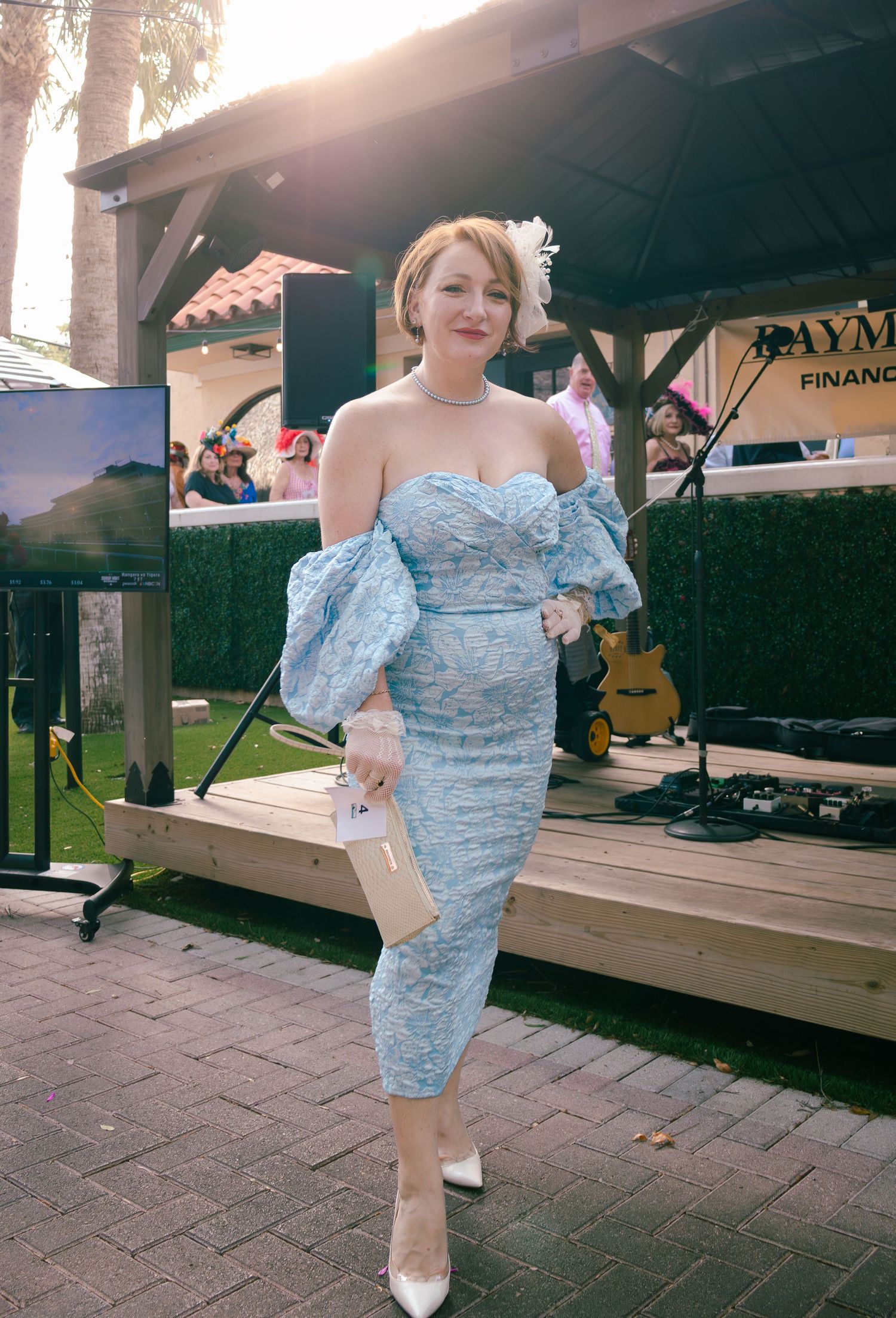 Woman in elegant blue off-shoulder lace dress with feathered hat at outdoor event.