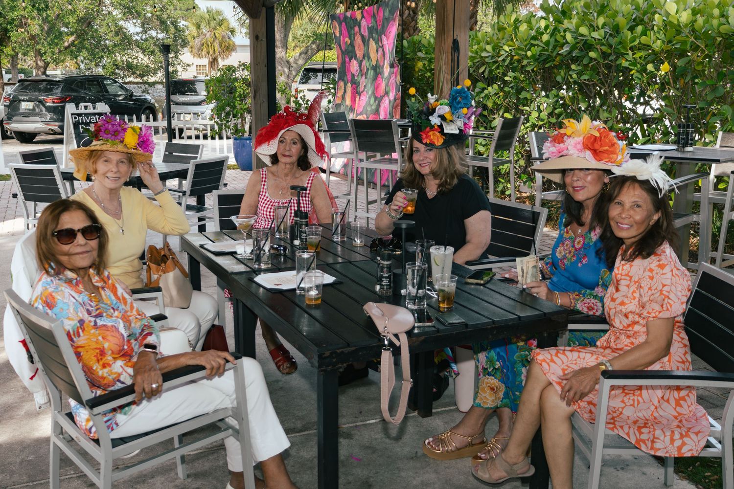 Six women wearing festive decorated hats pose together at an outdoor restaurant table during a Kentucky Derby party.