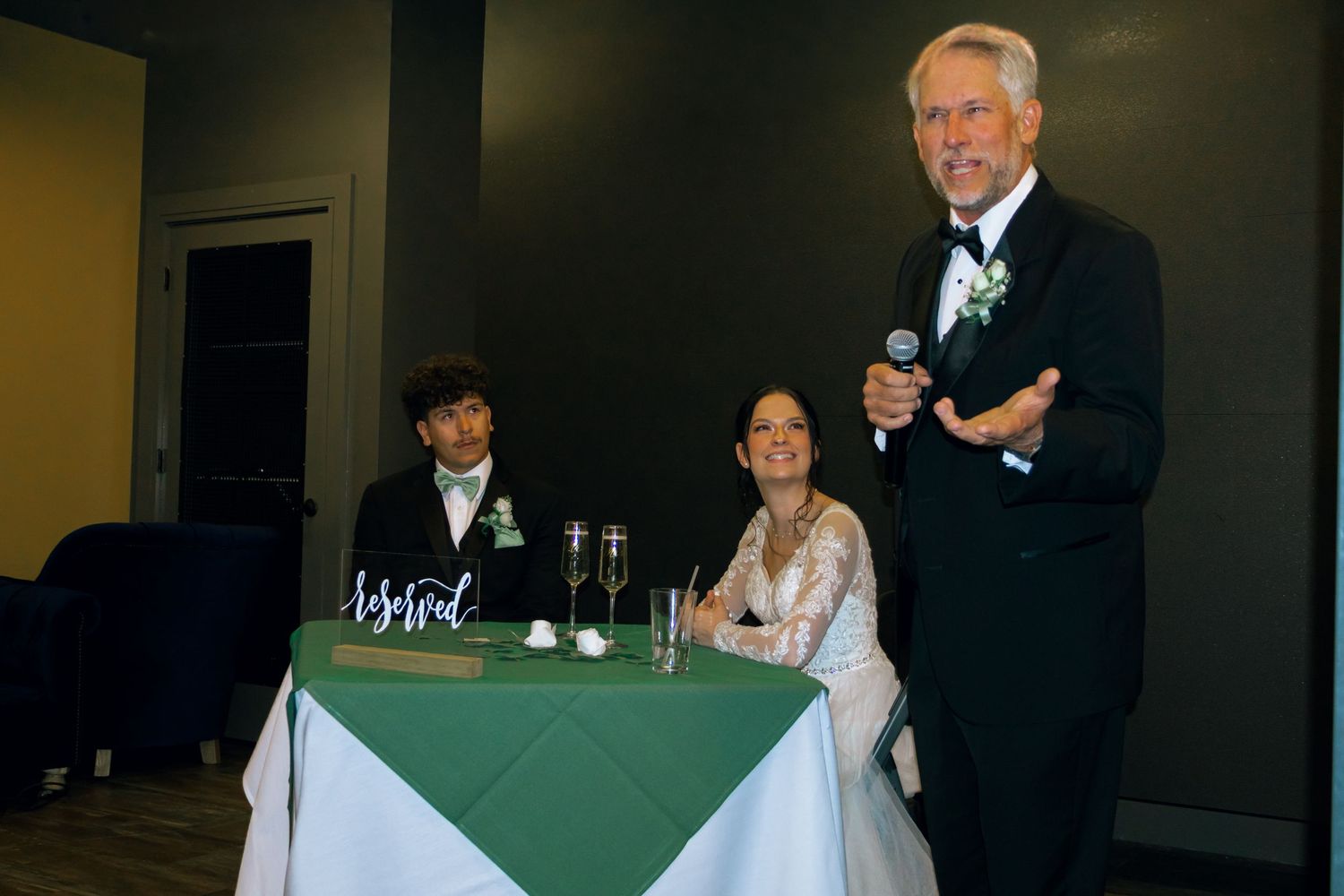 Father of bride giving wedding speech at sweetheart table with couple seated.