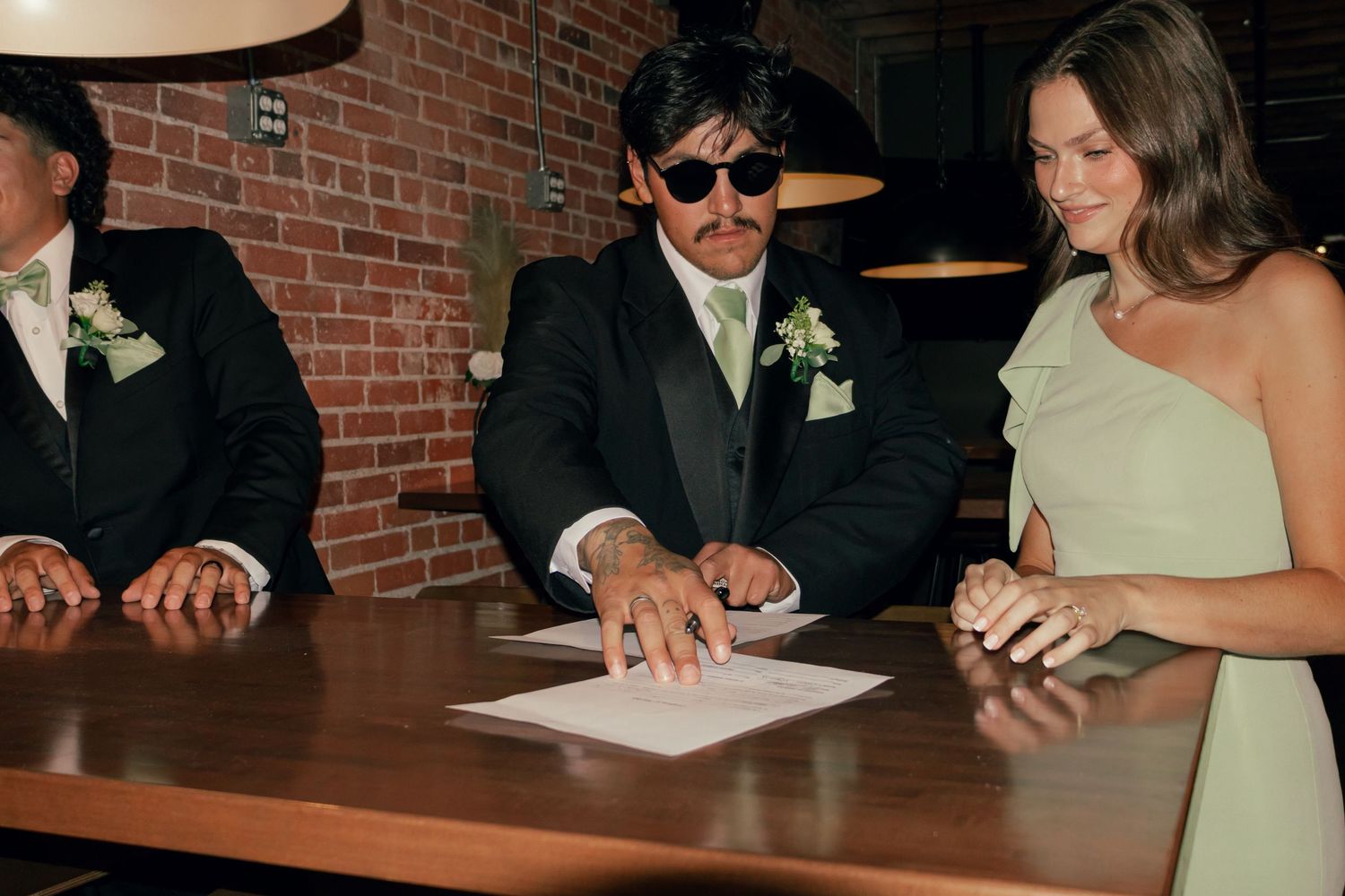 Wedding party signing marriage certificate at rustic brick wall venue.