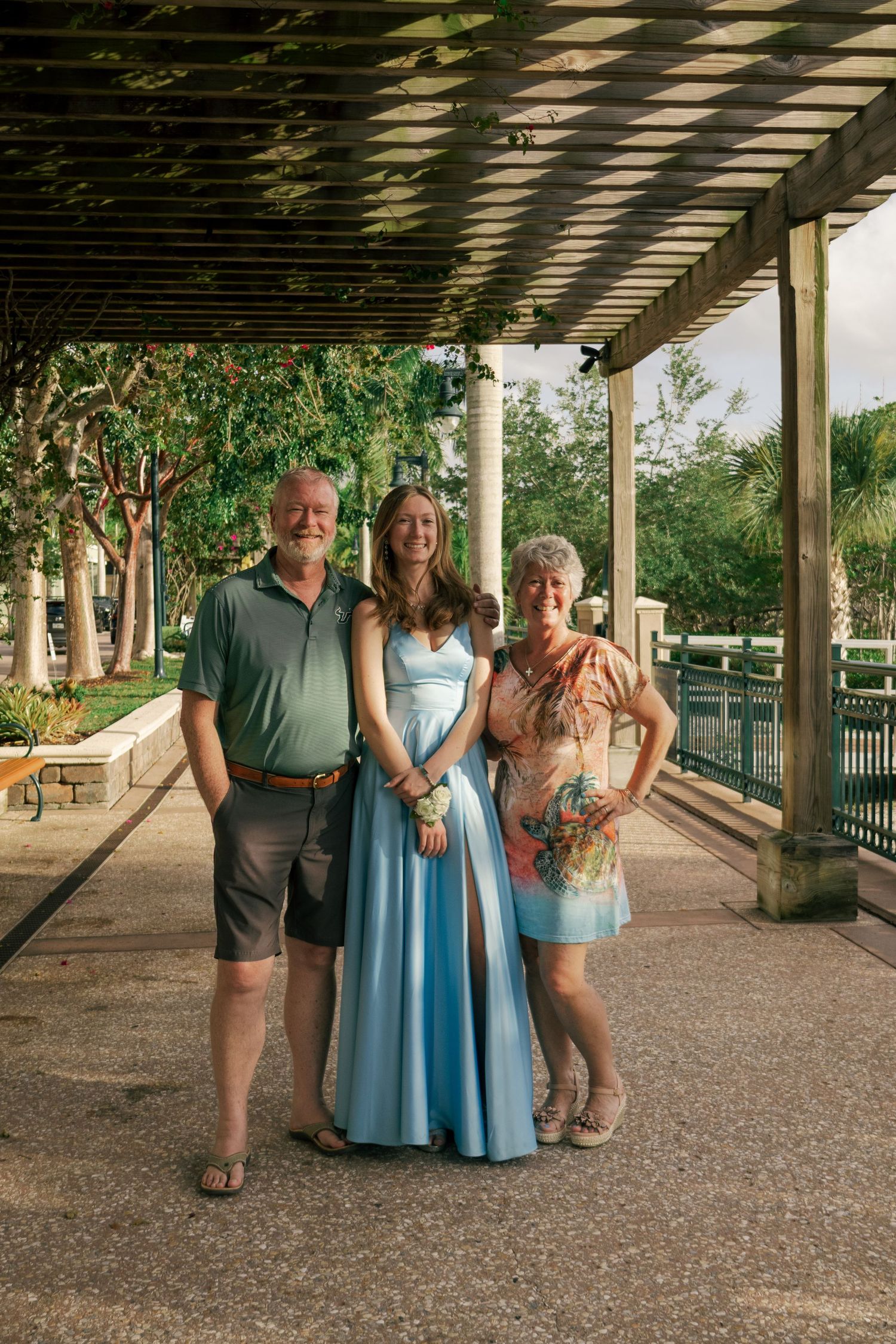 Young woman in a light blue formal gown posing with two older adults under a park pergola.