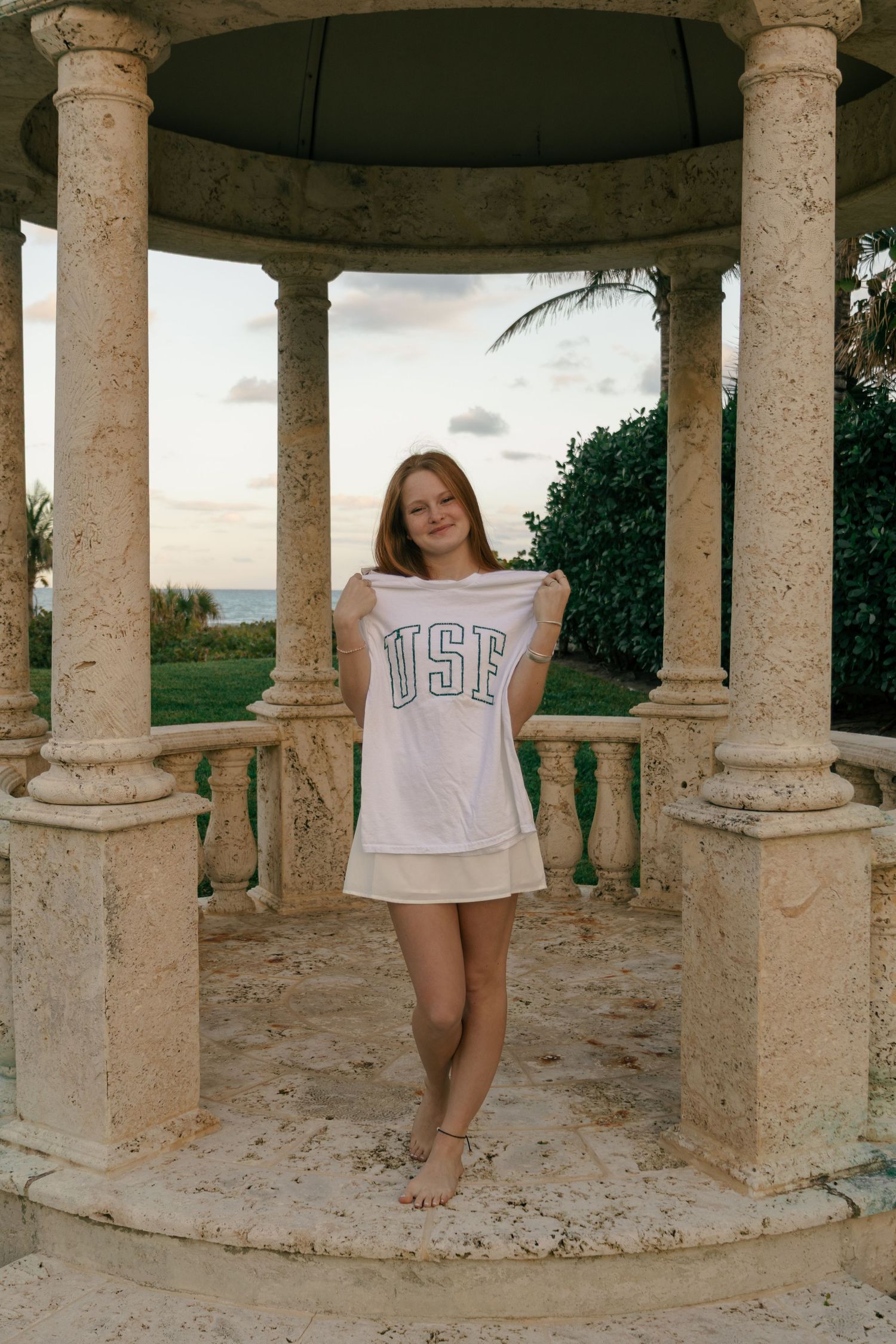 Smiling young woman holding USF college shirt standing barefoot in stone gazebo with ocean view background.