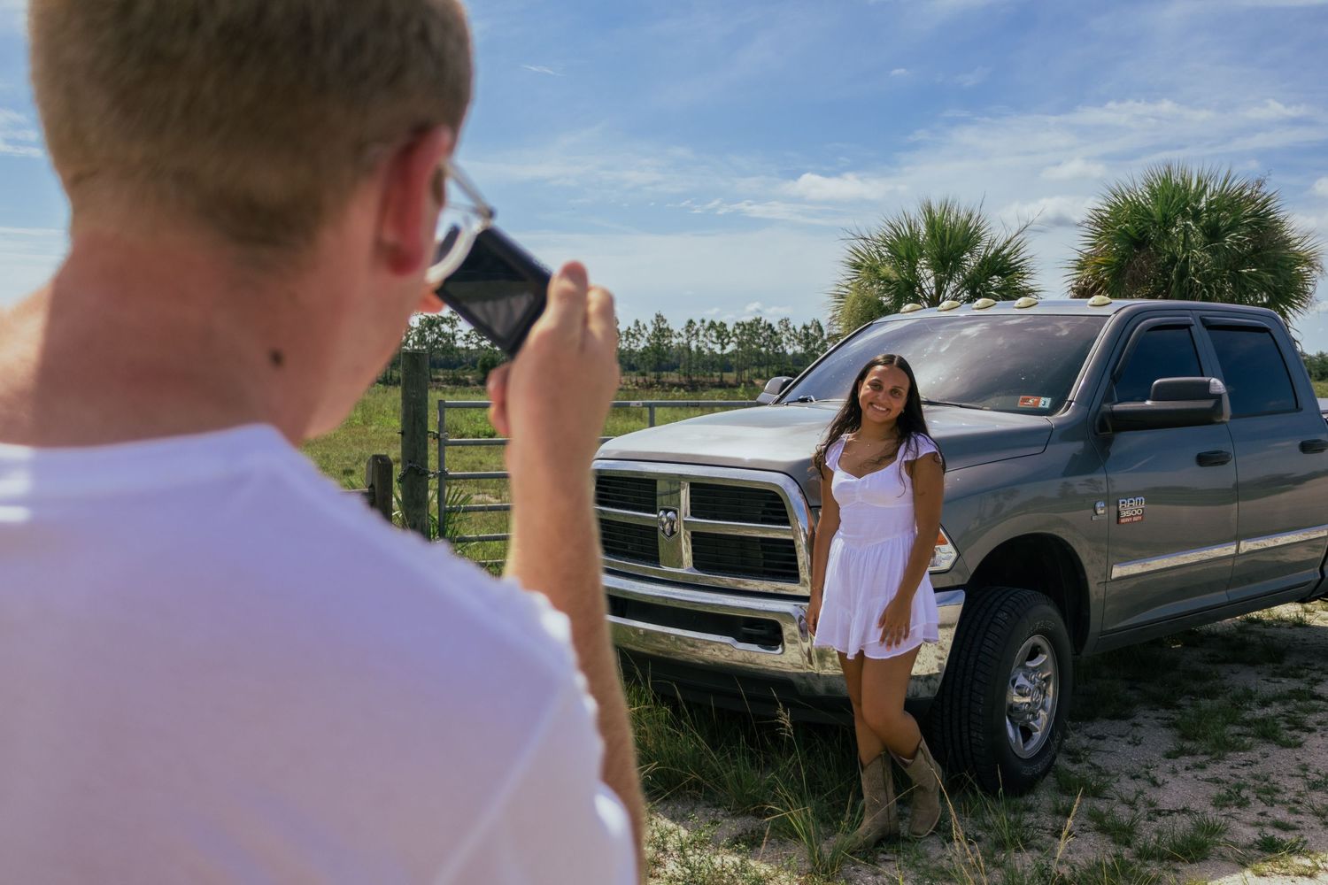 Young man photographing a woman in a white dress posing beside a gray Dodge Ram truck.