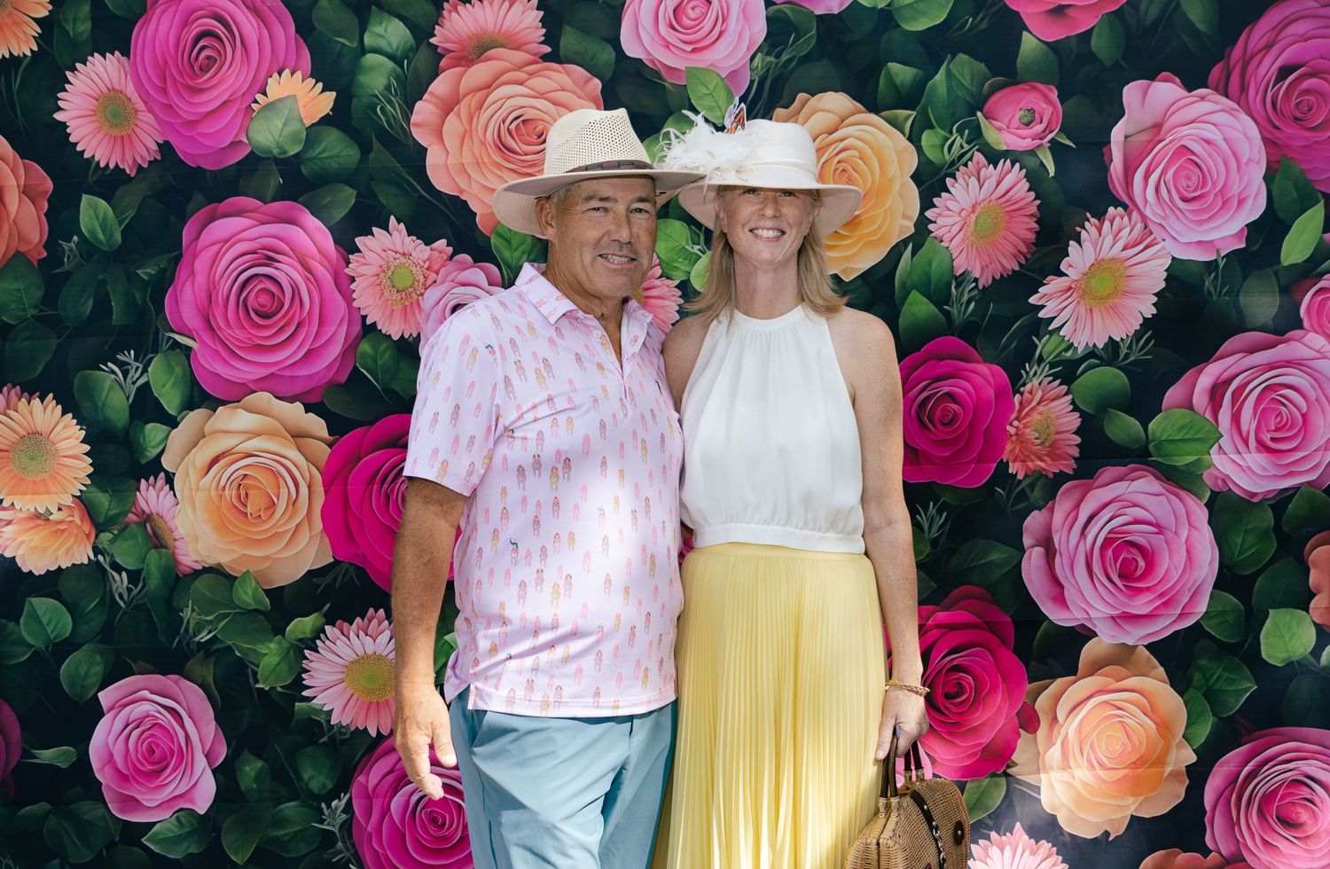 Smiling couple in summer attire posing together in front of a bright painted pink and orange floral rose backdrop.
