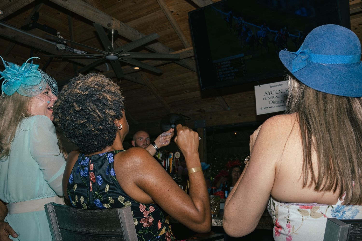 Women in elegant fascinators and wide-brim hats dance and socialize at a Kentucky Derby themed party.