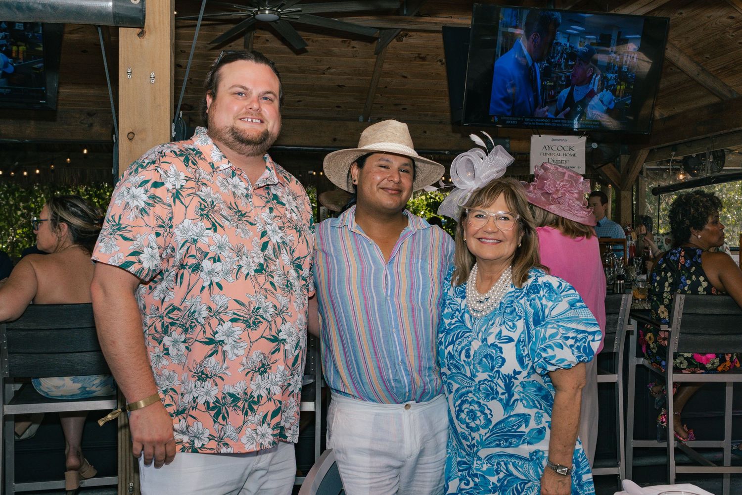 Three friends pose together at an outdoor bar, wearing colorful summer attire and festive hats.