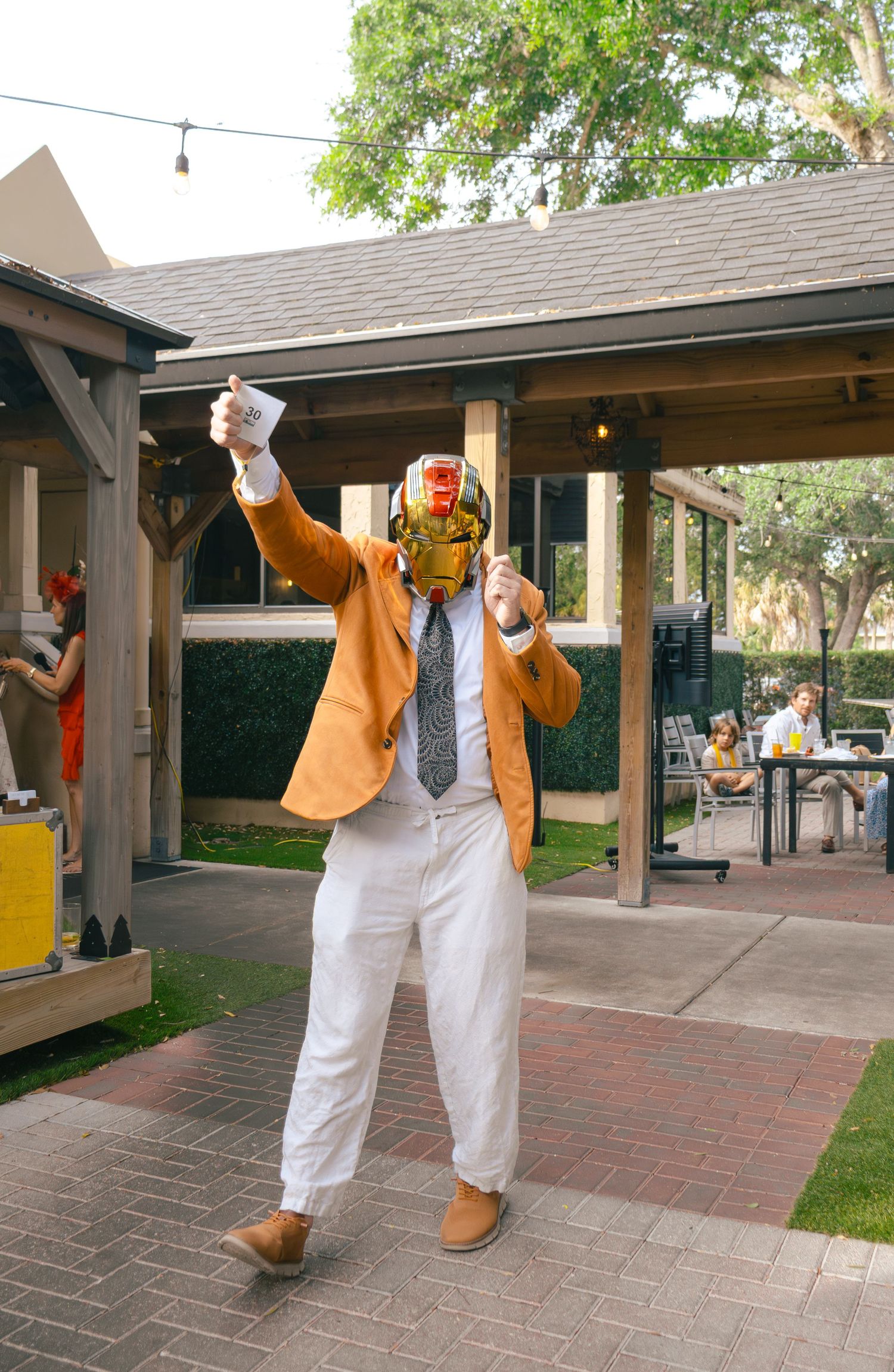 A performer in a gold helmet, orange blazer, and white pants strikes a celebratory pose at an outdoor event.