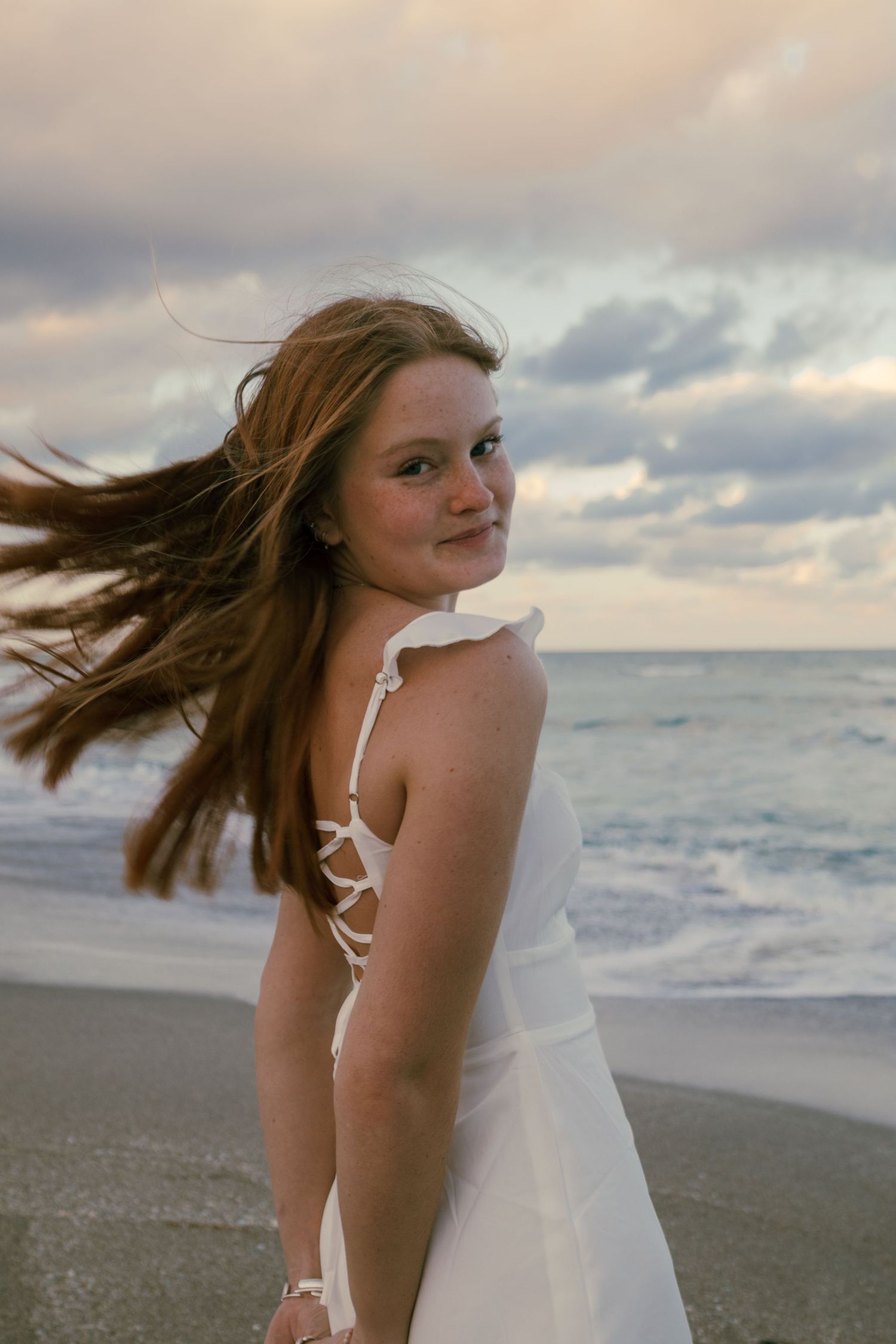 Young woman in white dress on beach at sunset with hair blowing in the wind.