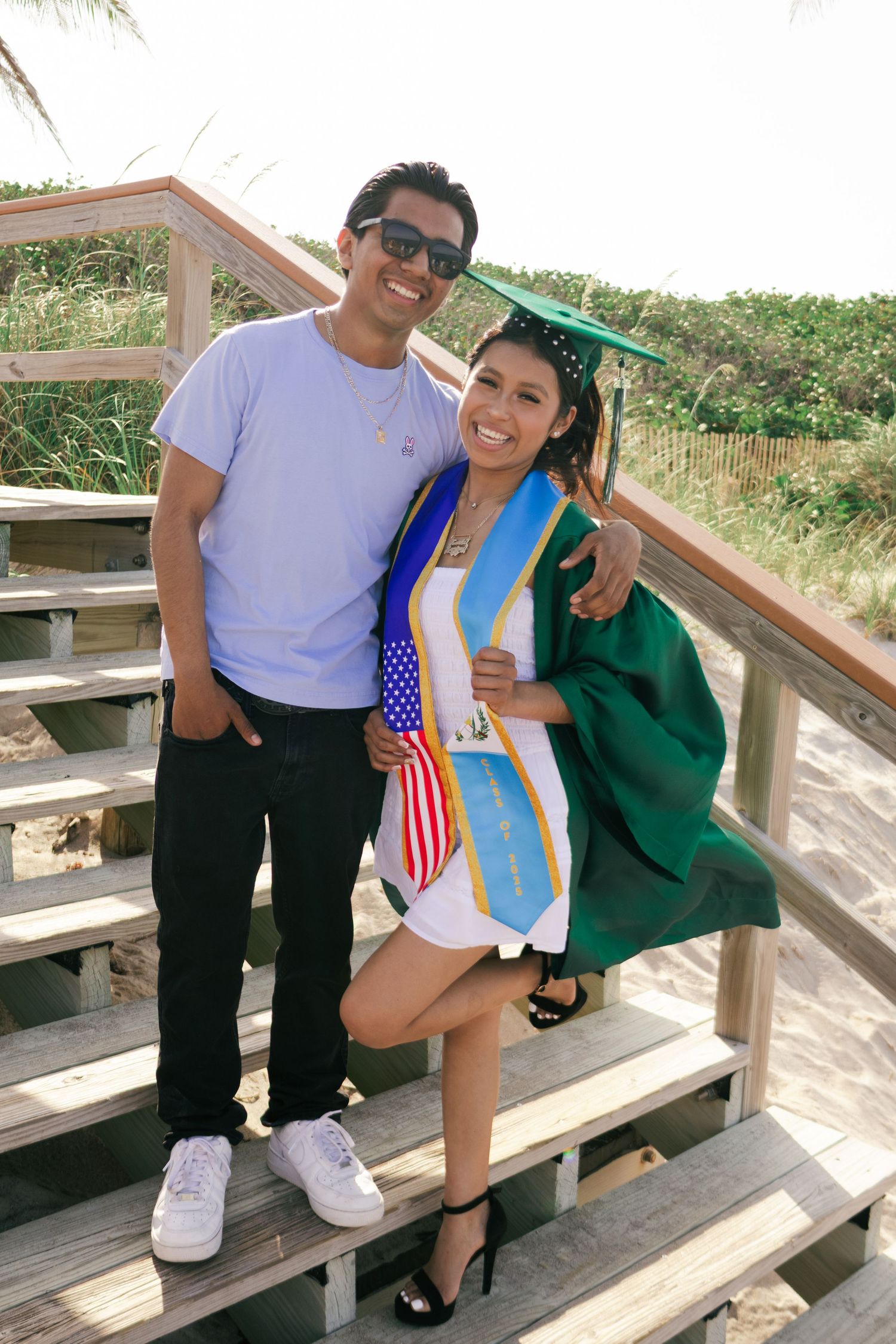 Smiling graduate in green cap and gown poses with brother on wooden beach boardwalk stairs in sunlight.