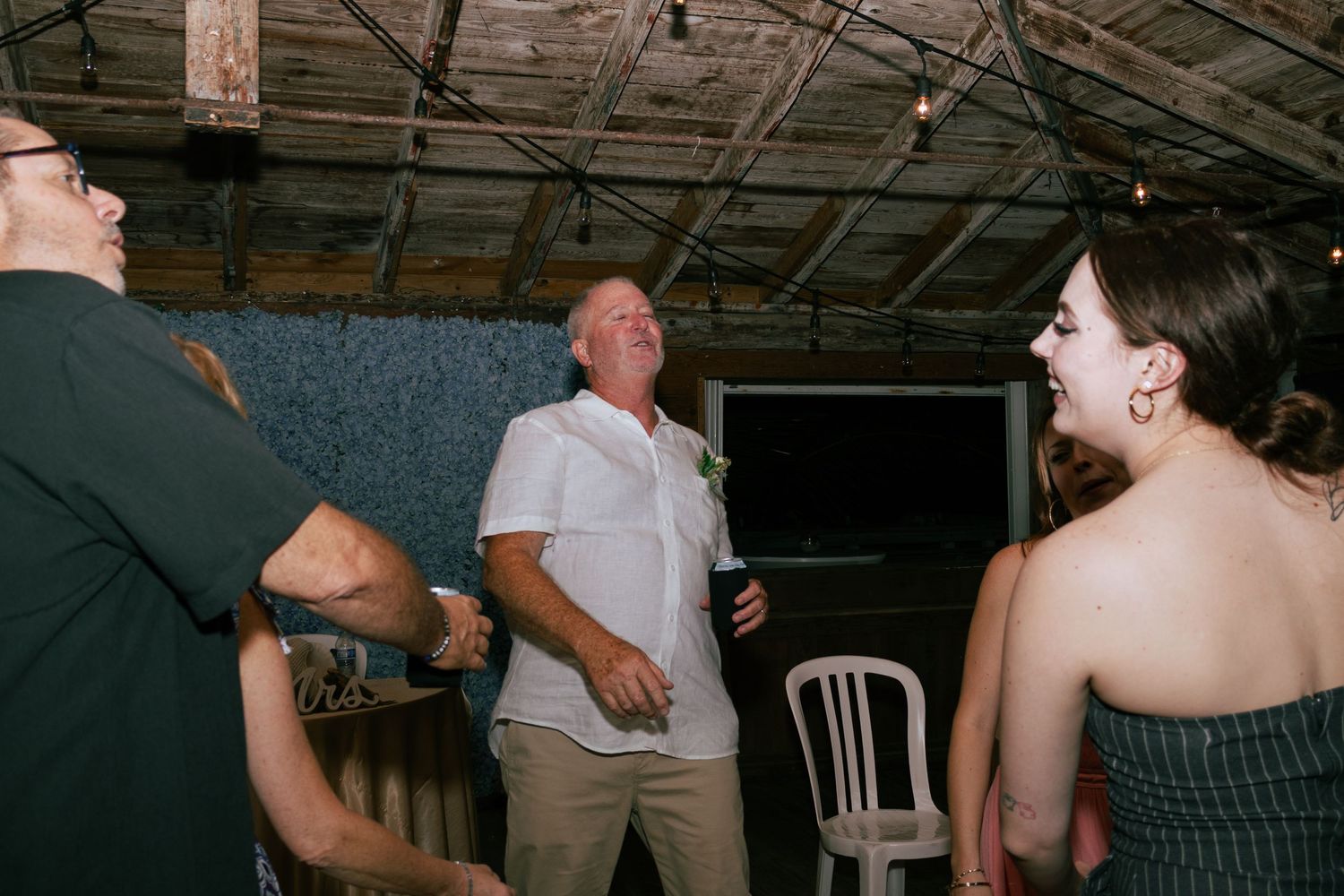 Man in white shirt laughing joyfully with guests at rustic wedding venue.