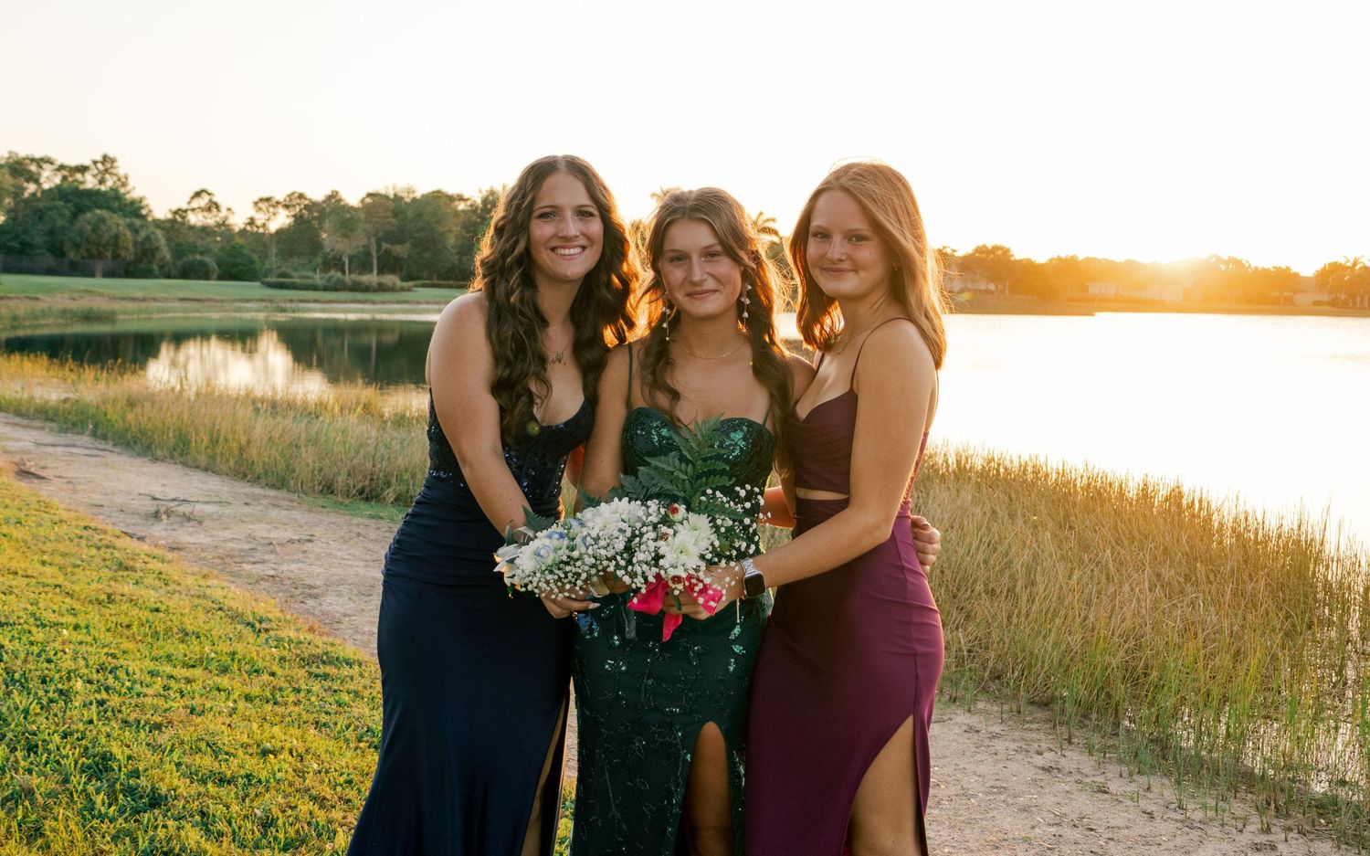 Three teenage girls in formal prom gowns pose together at sunset near a scenic lakeside path.