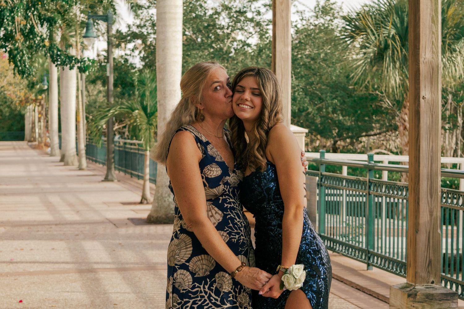 Two women in formal dresses sharing a sweet kiss on the cheek under a pergola walkway.