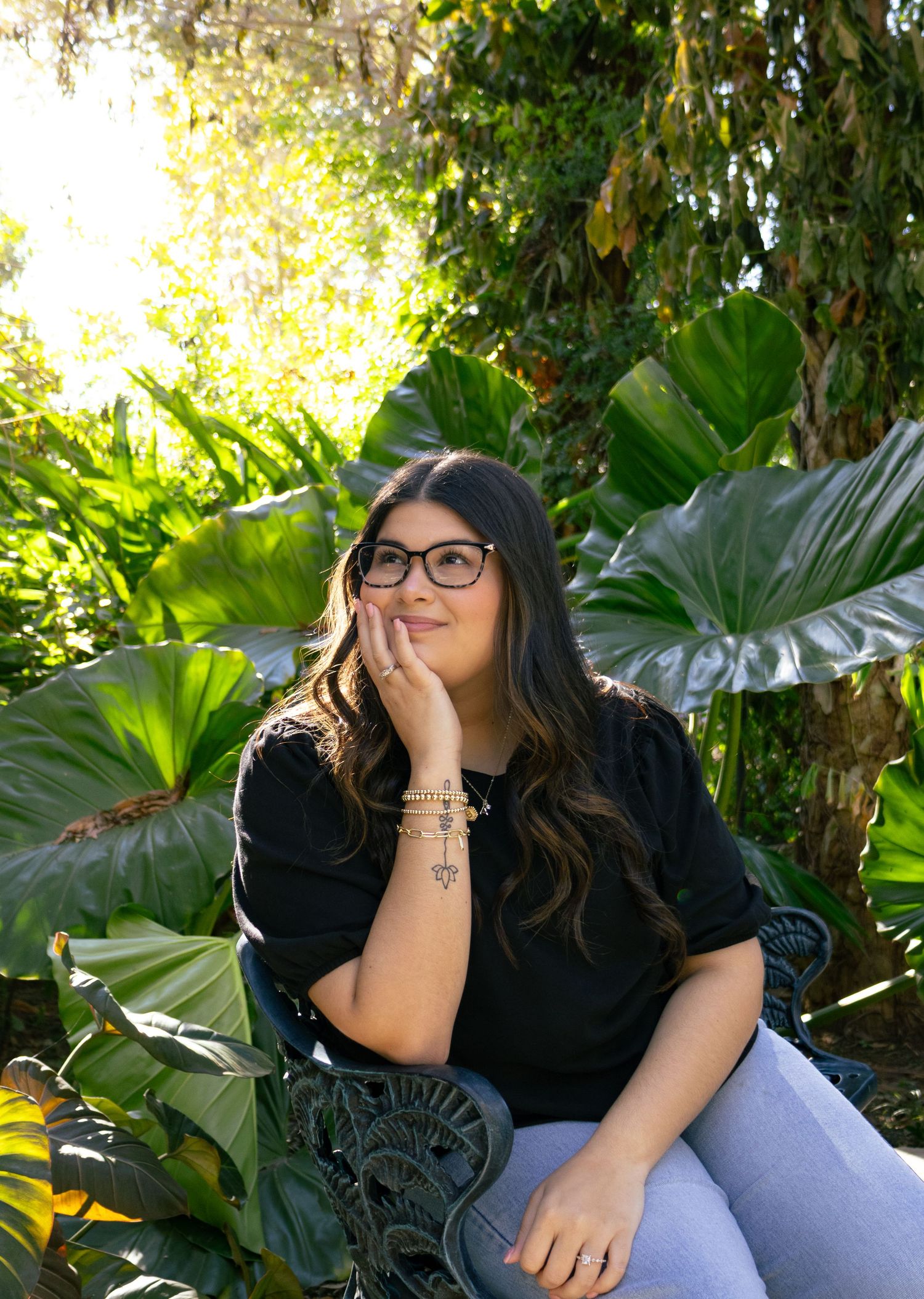 Woman with glasses gazes upward while sitting on iron bench surrounded by lush tropical garden foliage.