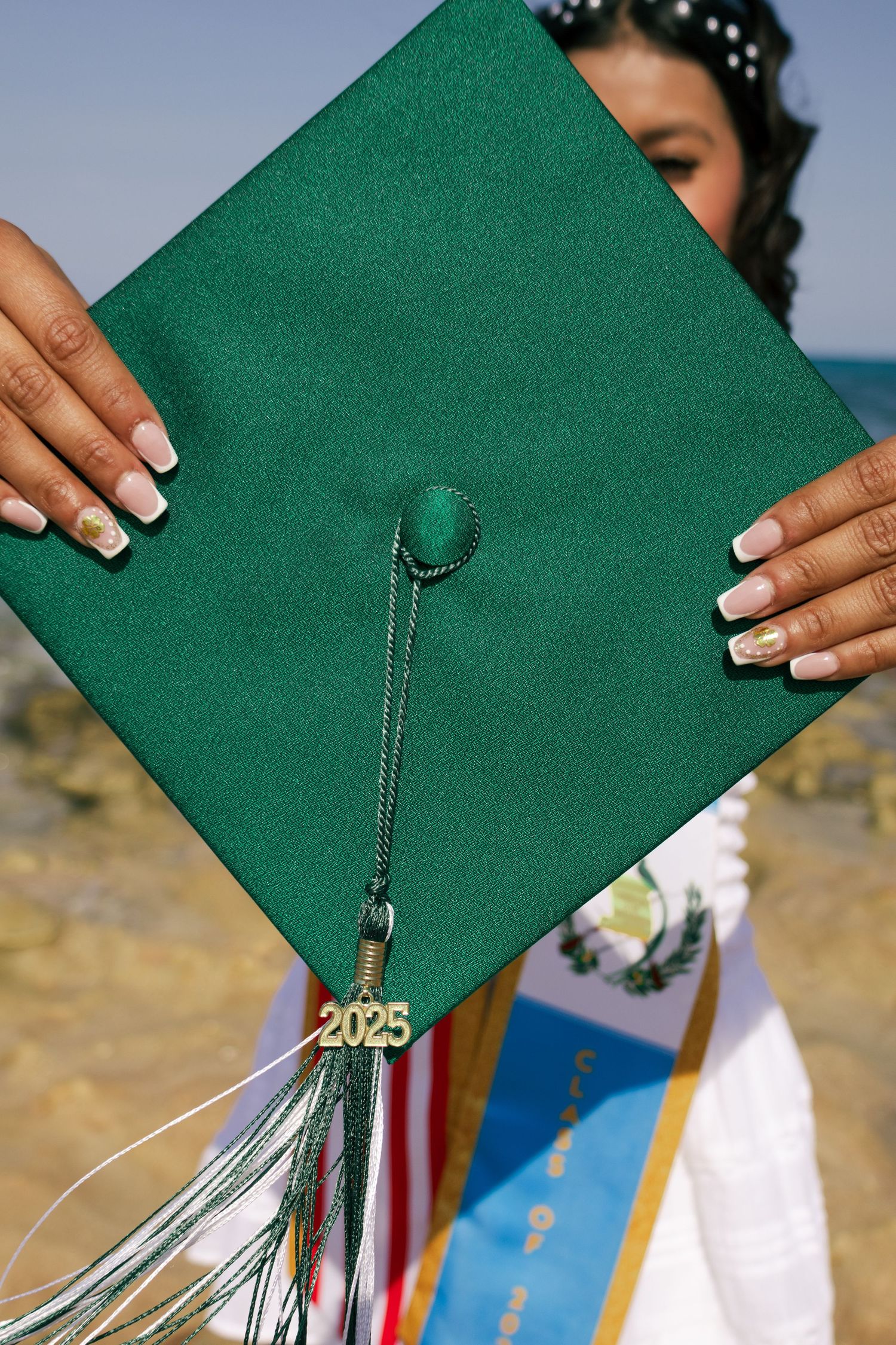 Close-up of green 2025 graduation cap with tassel held up by hands with French manicure.