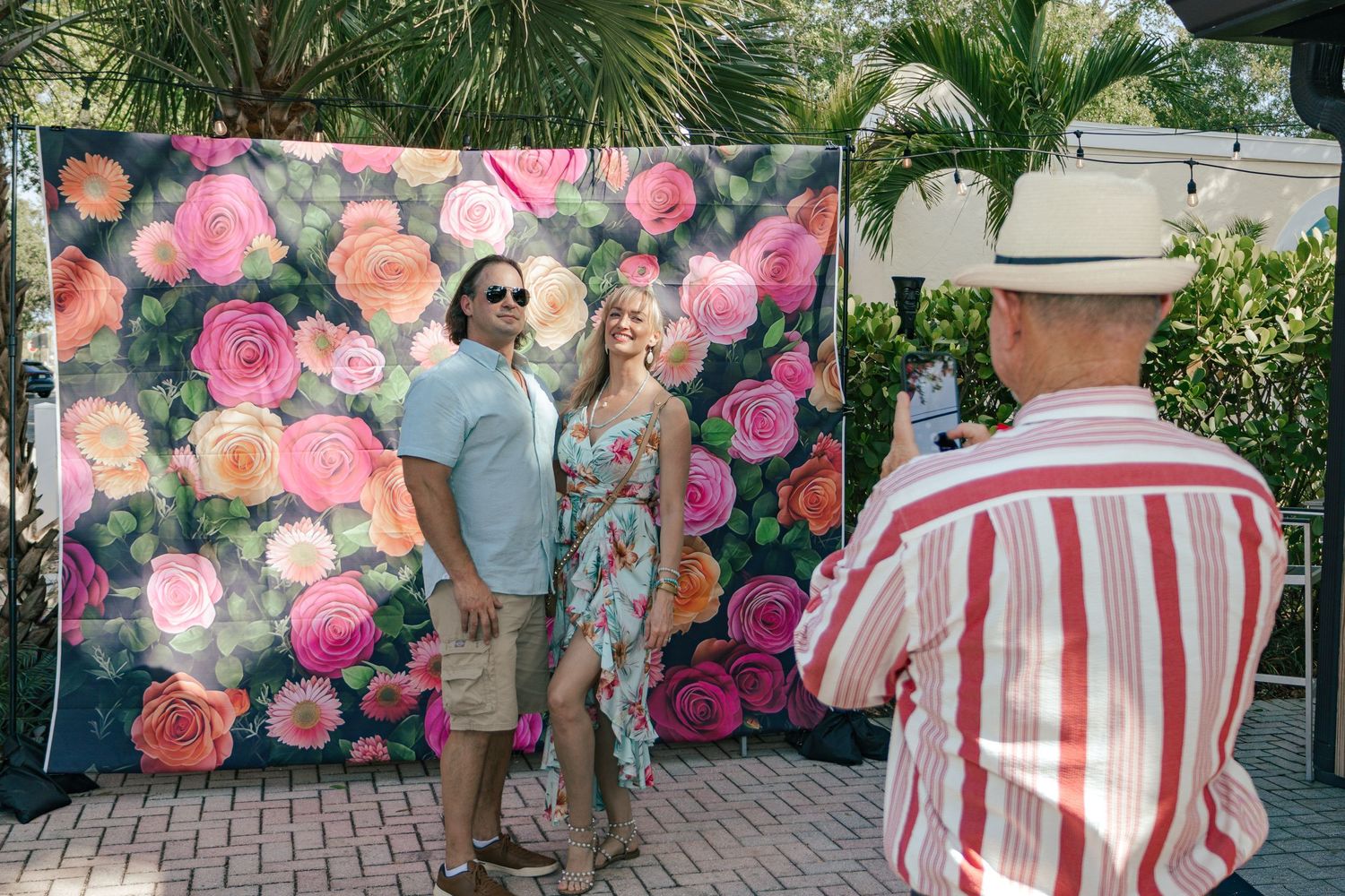 Couple posing for photo in front of colorful floral backdrop while man in striped shirt photographs them.