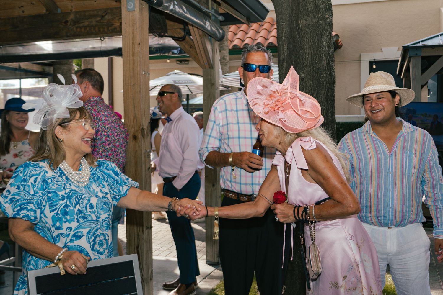 Women in festive hats shaking hands at an outdoor Kentucky Derby themed party event.