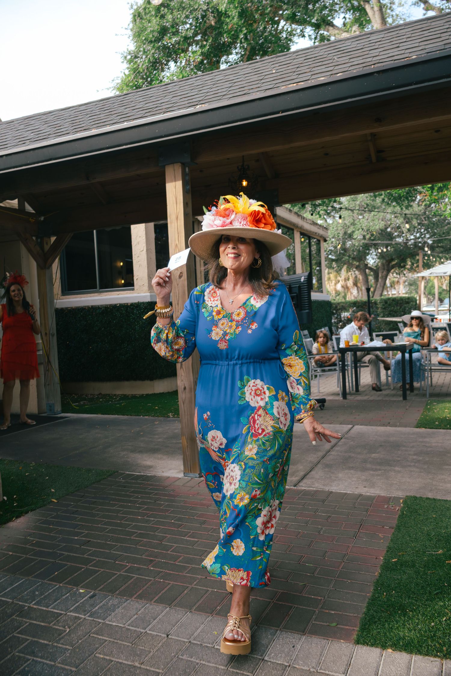 Woman in blue floral dress and decorated wide-brim hat walking confidently at outdoor event.
