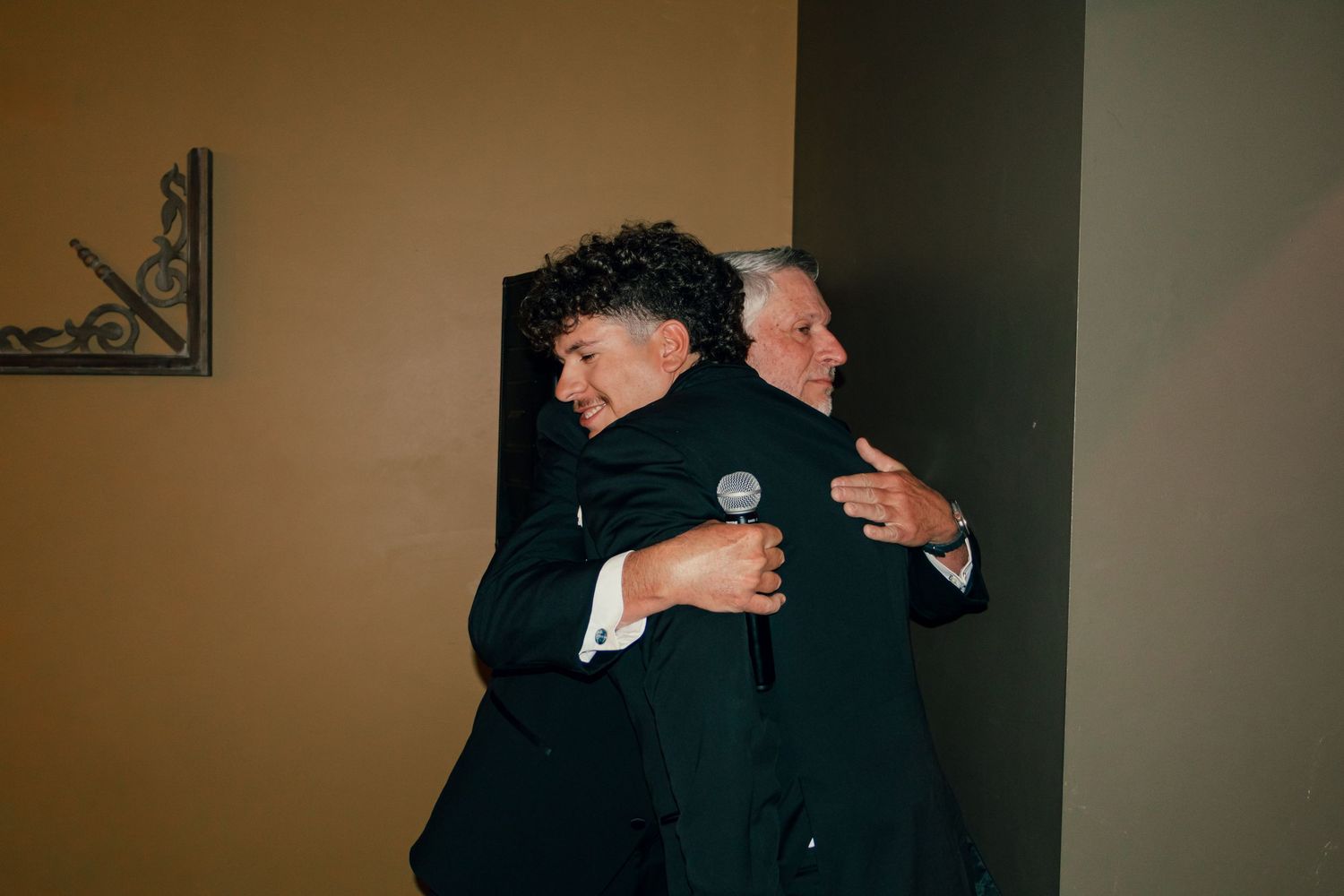 Two men in suits sharing a warm hug at a wedding reception.