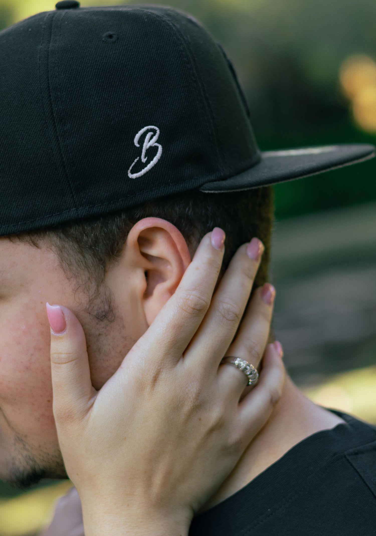 Close-up of a womans hand with pink nails and diamond ring gently cupping a mans face near his ear.