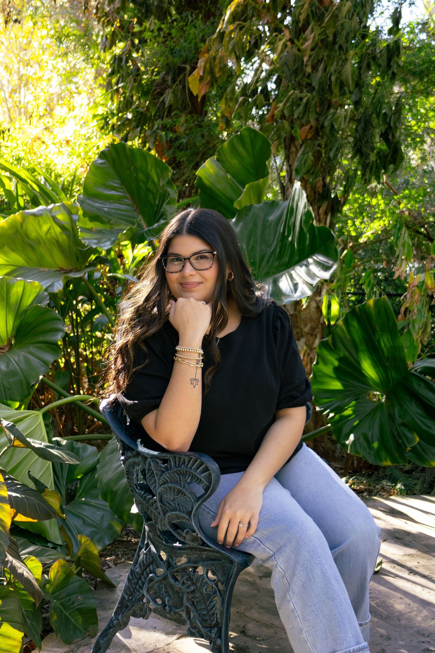 Woman with glasses and bracelets sits on ornate iron bench in sunlit garden with tropical plants.