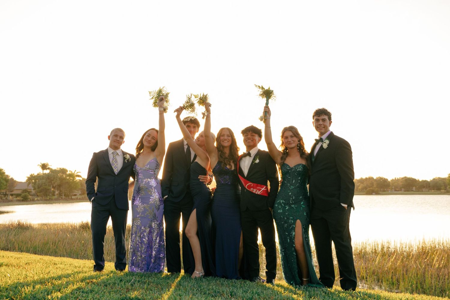 Seven prom students celebrate outdoors at golden hour near a lake, with girls raising bouquets.